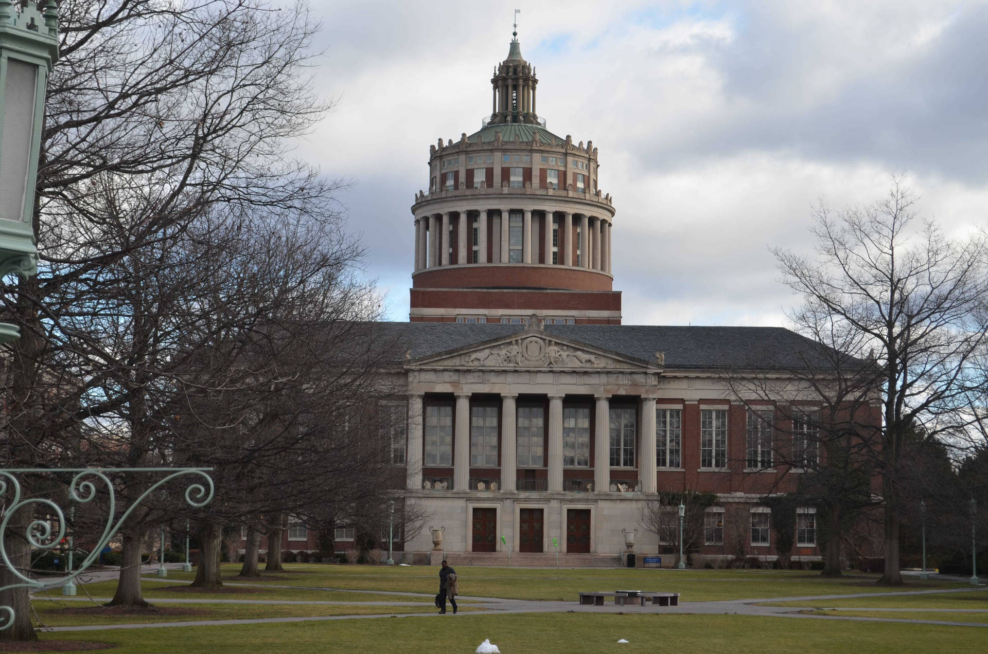  壁紙 — 一座宏偉的新古典主義大學建築，帶有圓頂塔樓，矗立在多雲的天空下，周圍是光禿禿的樹木和一片草地，草地上有一個孤獨的行人。