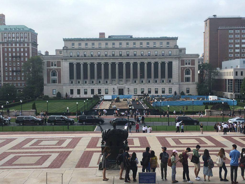 Library 桌布 壁紙 — 一座宏偉的新古典主義大學建築，帶有柱廊，矗立在一個圖案廣場後面，廣場上滿是學生和停泊的汽車，天空多雲。