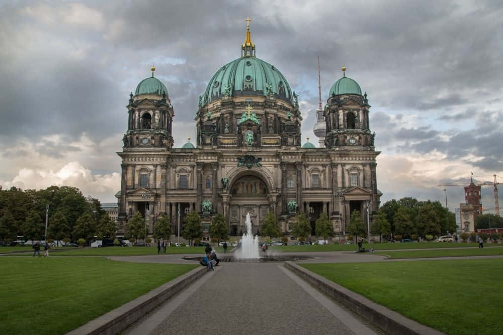 Berlin 桌布 壁紙 — 一座宏偉的新古典主義大教堂，擁有綠色圓頂，矗立在多雲的天空下，中央噴泉和修剪整齊的草坪構成畫框。