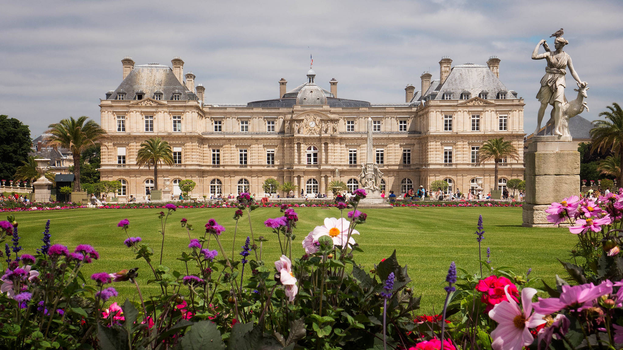 France 桌布 壁紙 — 一座宏偉的新古典主義宮殿，擁有華麗的建築，矗立在修剪整齊的草坪和充滿活力的花壇後面，天空多雲。