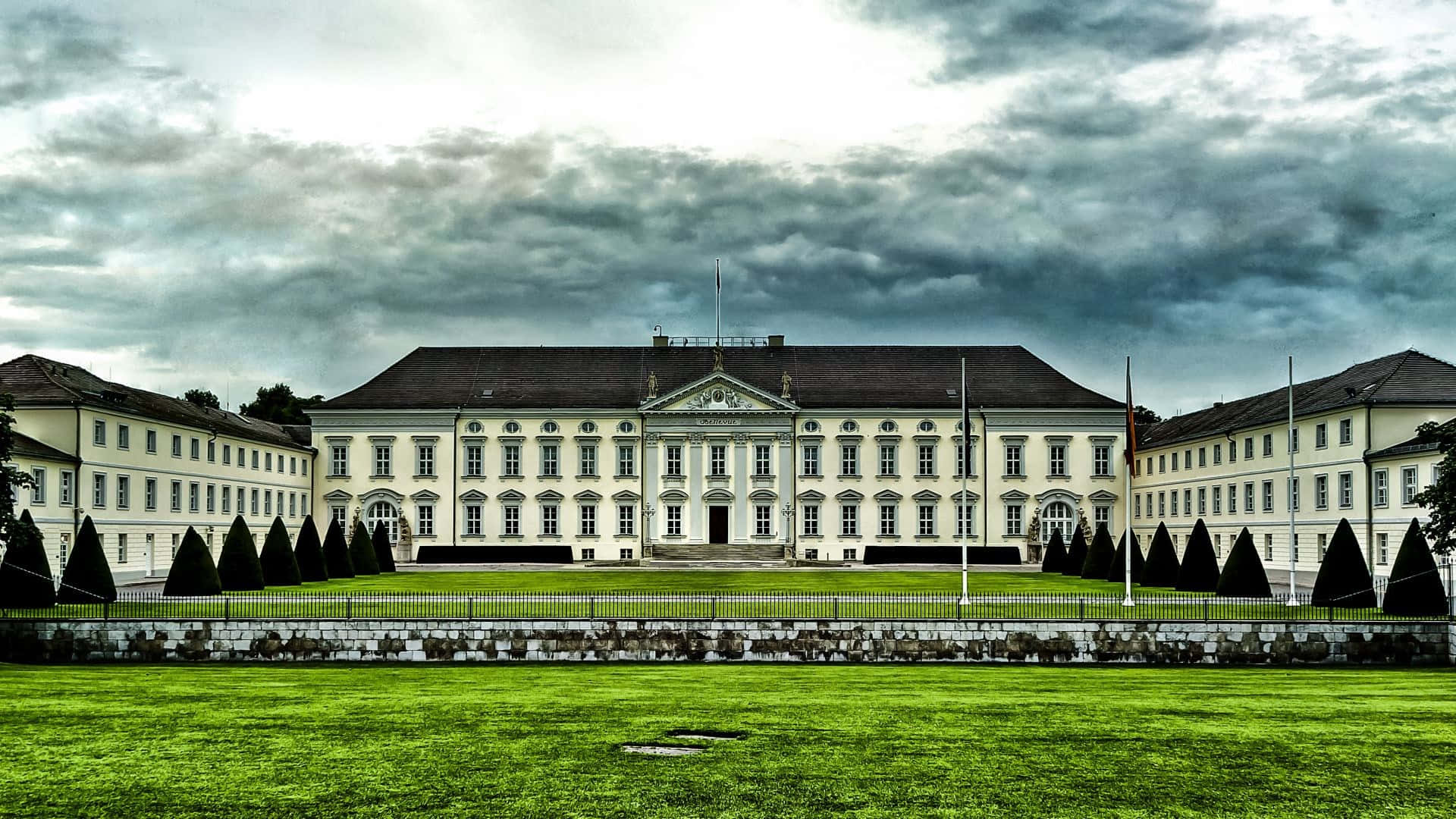 Berlin 桌布 壁紙 — 一座宏偉的新古典主義宮殿，白色牆壁和深色屋頂，矗立在戲劇性的多雲天空下，兩側對稱的翼樓和充滿活力的綠色草坪。