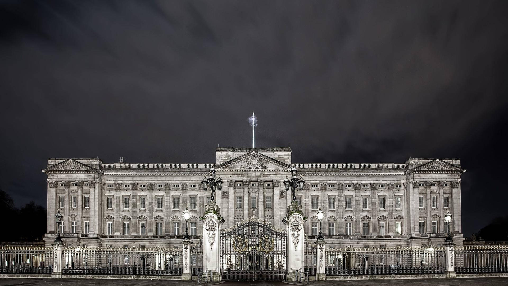 England 桌布 壁紙 — 一座宏偉的新古典主義宮殿在夜間被照亮，天空陰暗多雲，頂上飄揚著旗幟。