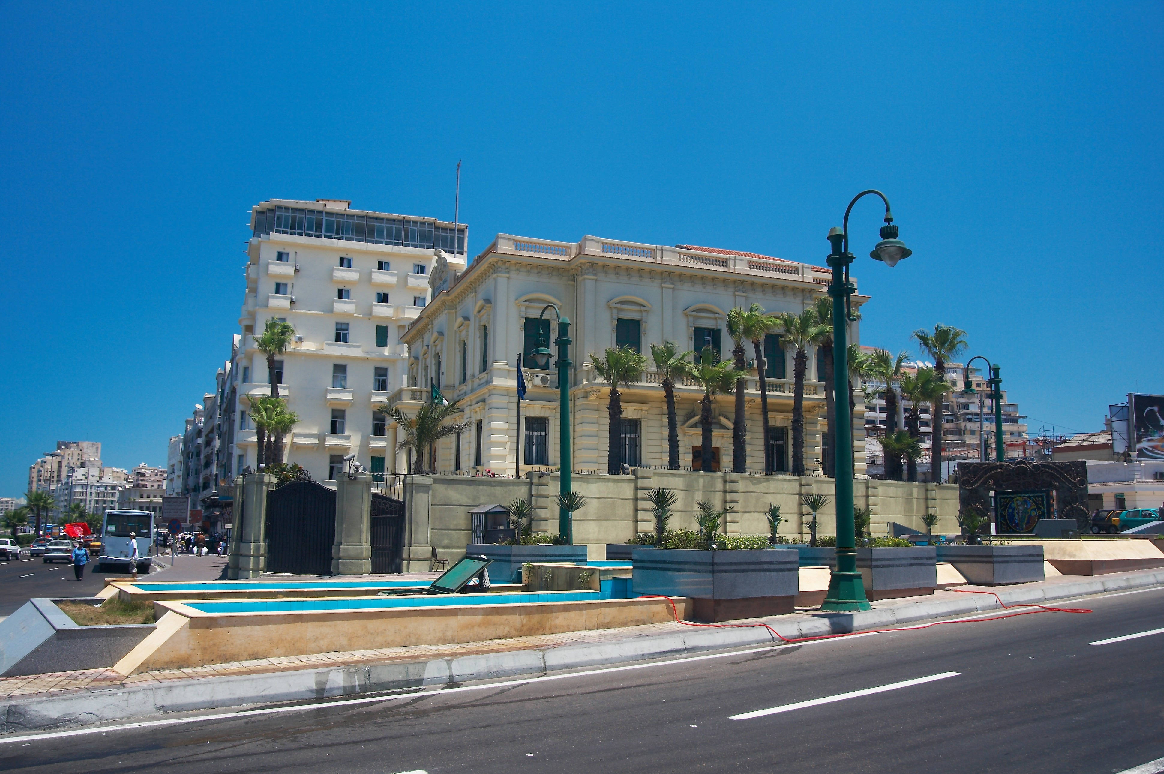 Alexandria 桌布 壁紙 — 一座宏偉的新古典主義建築，周圍有棕櫚樹，矗立在晴朗的藍天下，旁邊是一條有綠色路燈的現代街道。