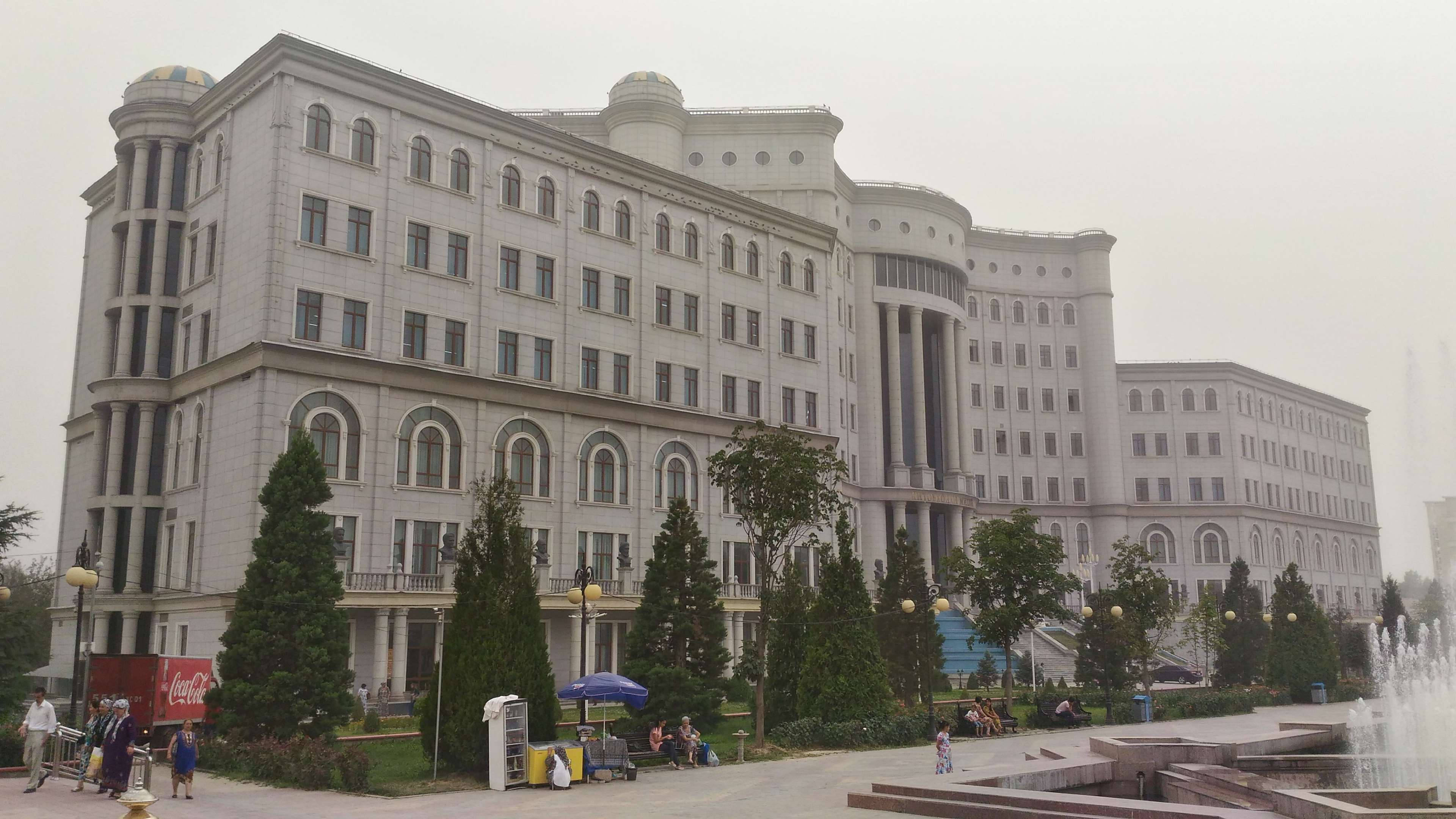 Tajikistan 桌布 壁紙 — 一座宏偉的新古典主義建築，擁有拱形窗戶和圓頂，矗立在修剪整齊的樹木和噴泉後面，位於陰天下的公共廣場。