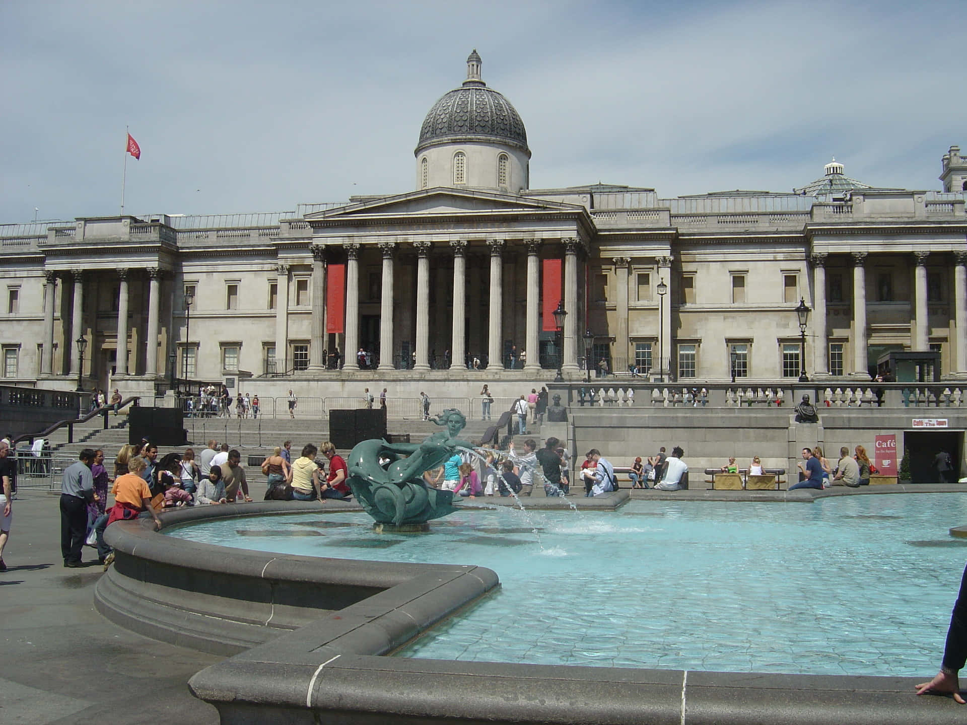 Museum 桌布 壁紙 — 一座宏偉的新古典主義建築，擁有柱子和圓頂，俯瞰著熙熙攘攘的噴泉，人們在陽光下放鬆和玩耍。