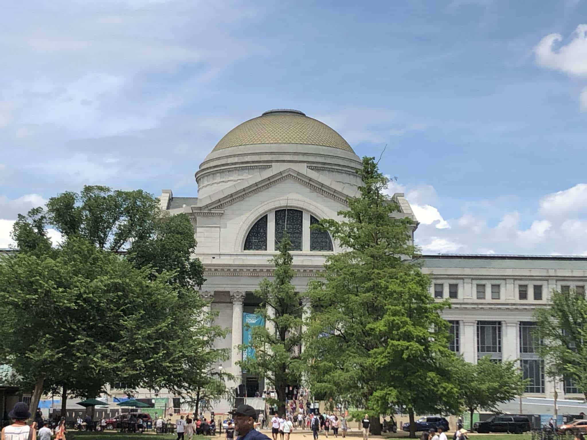 history 桌布 壁紙 — 一座宏偉的新古典主義建築，擁有金色圓頂和白色柱子，矗立在明亮的藍天下，周圍環繞著茂密的綠樹和熙熙攘攘的人群。