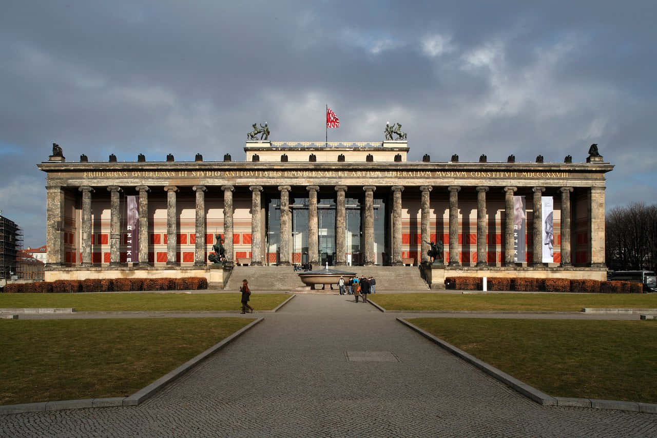 Berlin 桌布 壁紙 — 一座宏偉的新古典主義建築，擁有高大的柱子和雕像，在陰天天空下，通過寬闊的石徑到達，遊客稀少。