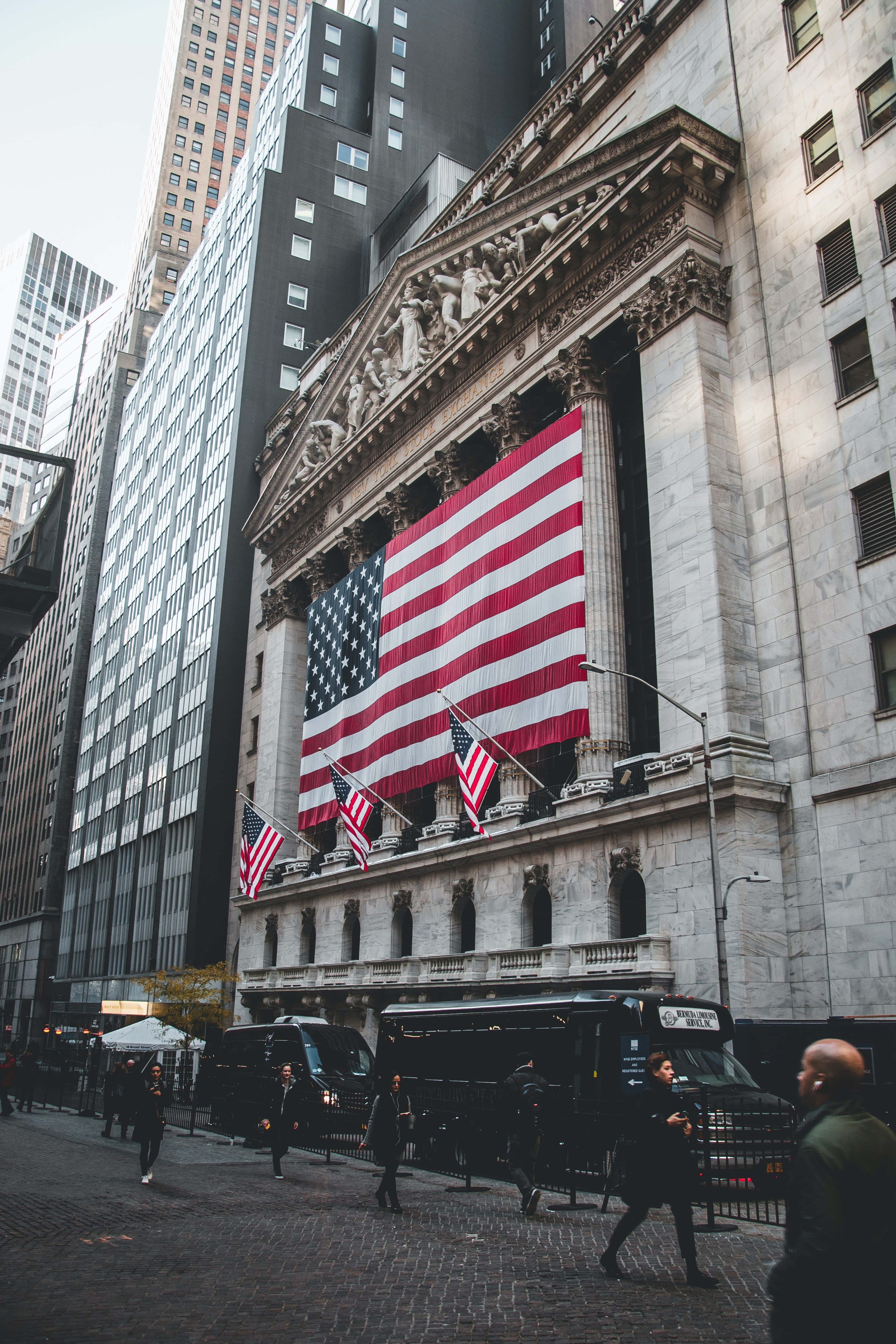 Wall Street 桌布 壁紙 — 一座宏偉的新古典主義建築，正面懸掛著巨大的美國國旗，矗立在繁華的城市街道上。