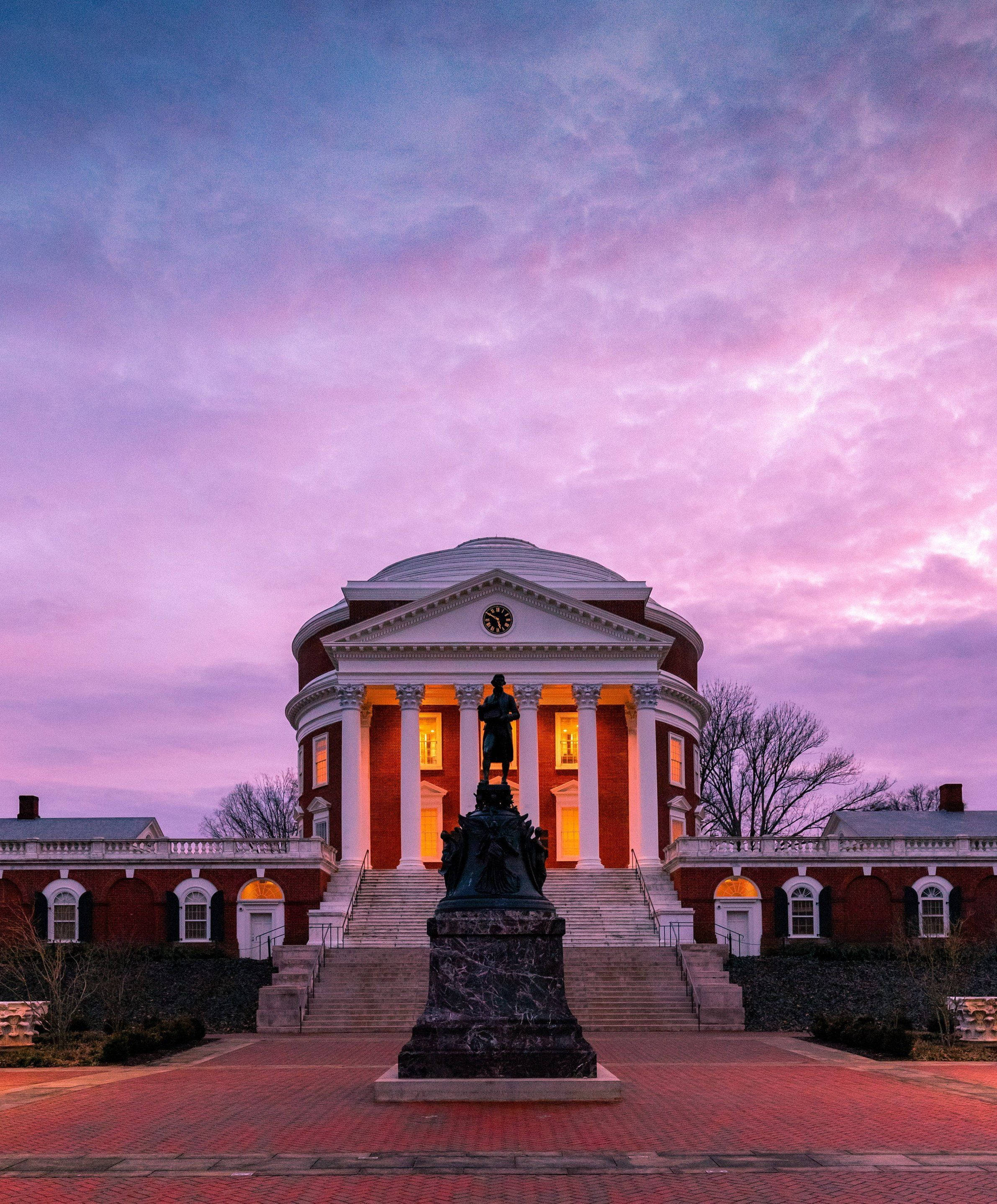 Virginia 桌布 壁紙 — 一座宏偉的新古典主義建築，窗戶發光，矗立在充滿活力的粉紅色和紫色暮色天空下，由大理石基座上的雕像框住。