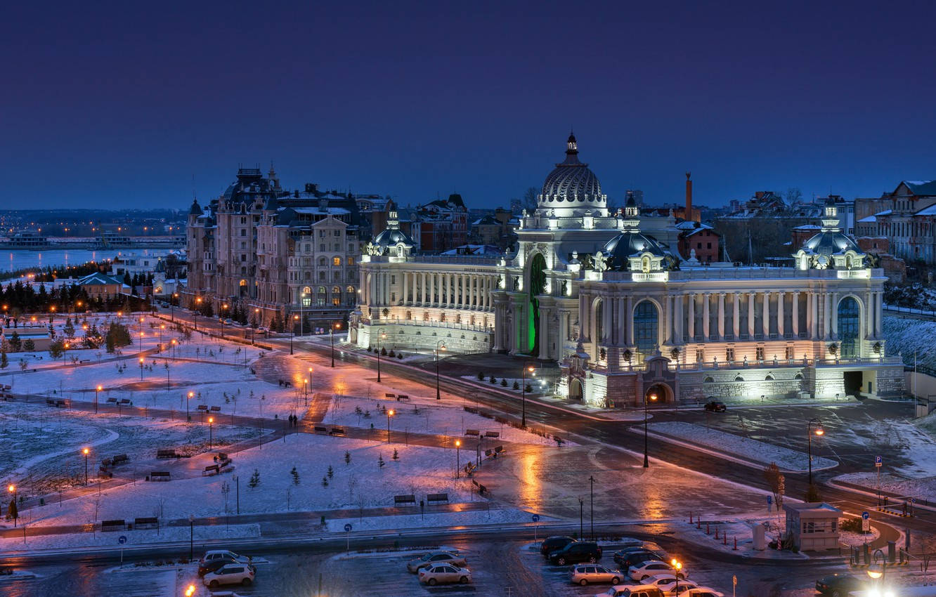 Kazan 桌布 壁紙 — 一座宏偉的新古典主義建築，閃爍的燈光在暮色中莊嚴地矗立在白雪覆蓋的城市廣場上。