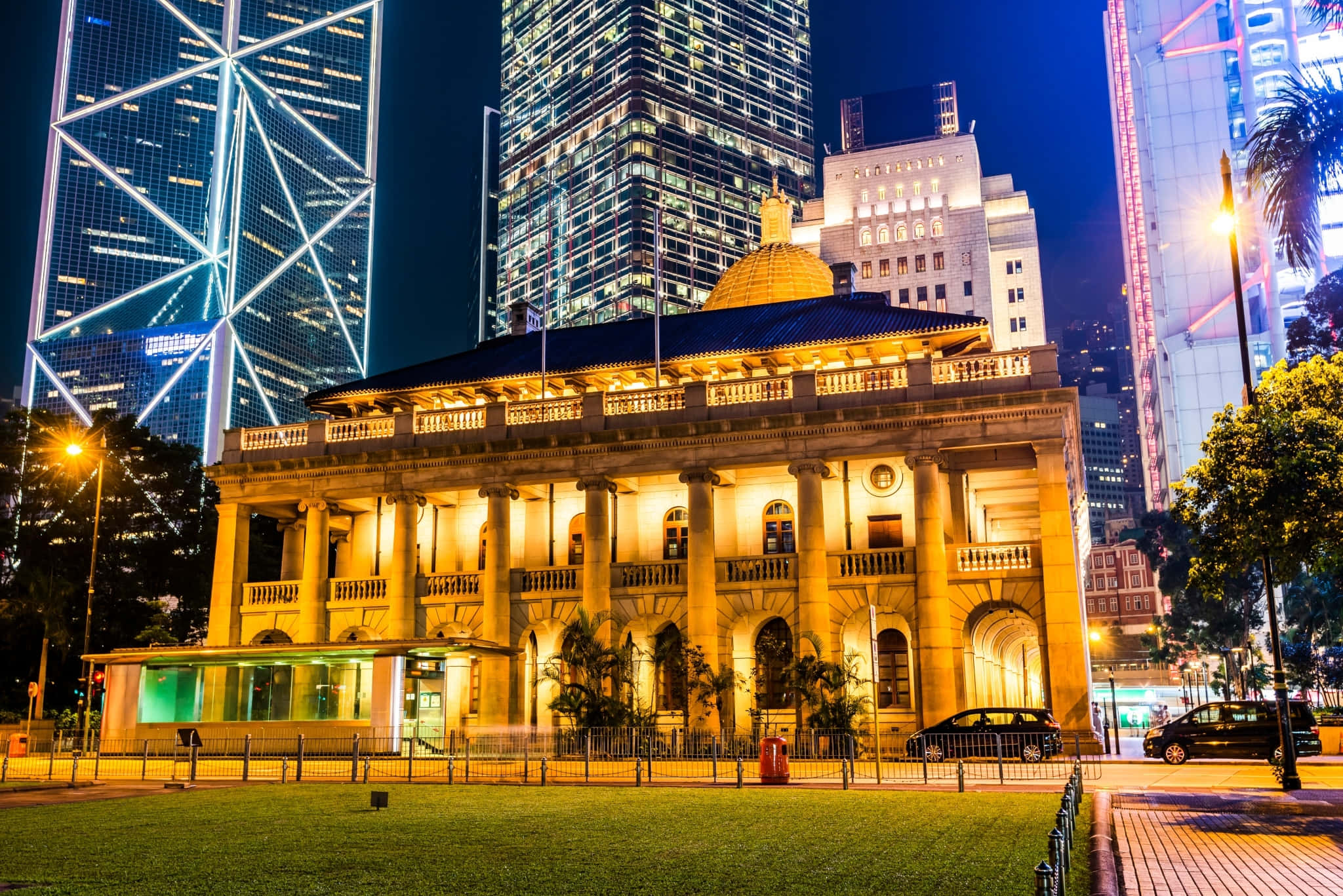 Beautiful Architecture 桌布 壁紙 — 一座宏偉的新古典主義建築在夜晚溫暖地發光，被高聳的照明摩天大樓和茂密的綠植所環繞。