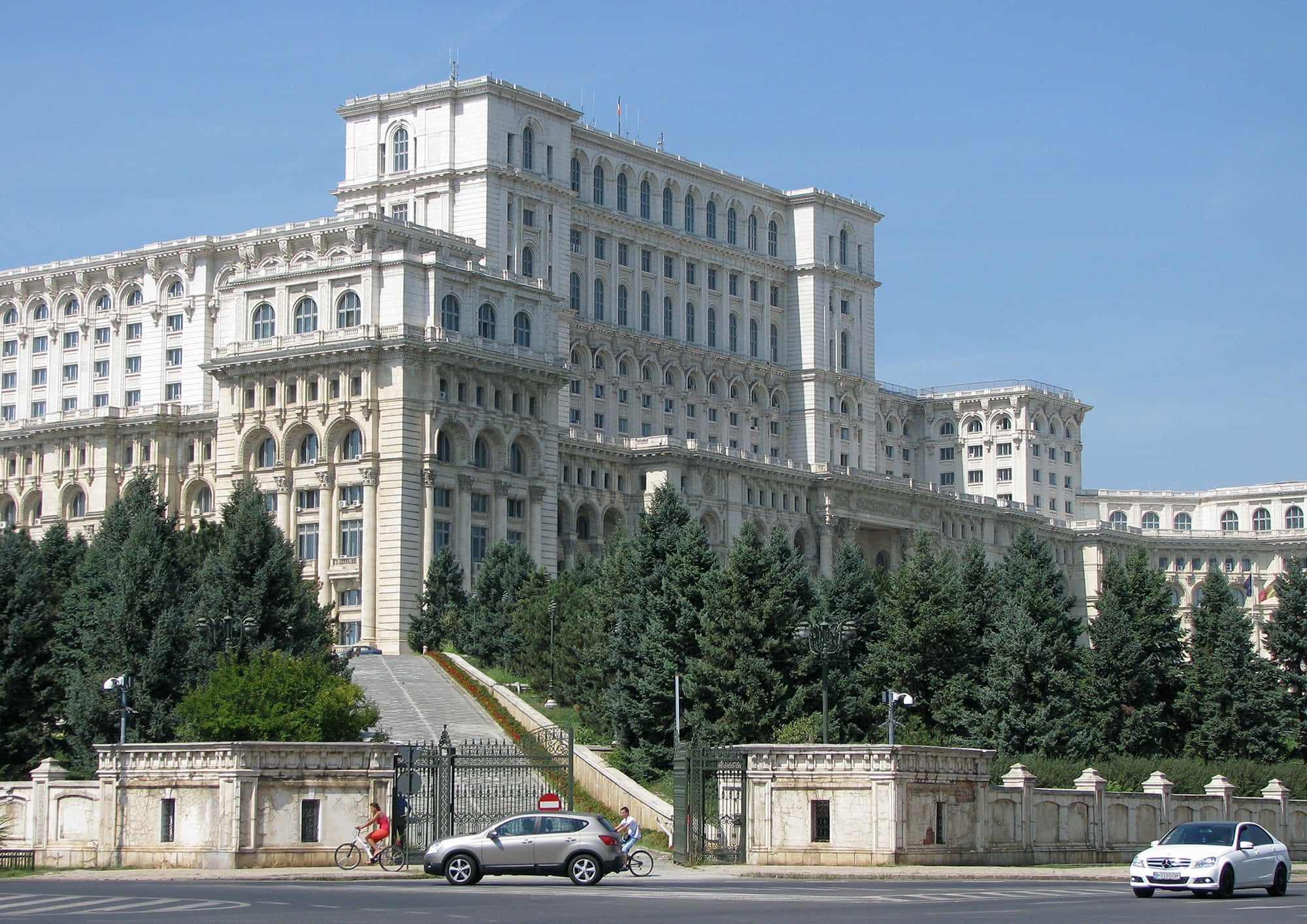 高清 桌布 壁紙 — 一座宏偉的新古典主義政府建築，白色石砌外牆和拱形窗戶，在晴朗的藍天下，周圍有常綠樹木和行駛的汽車。