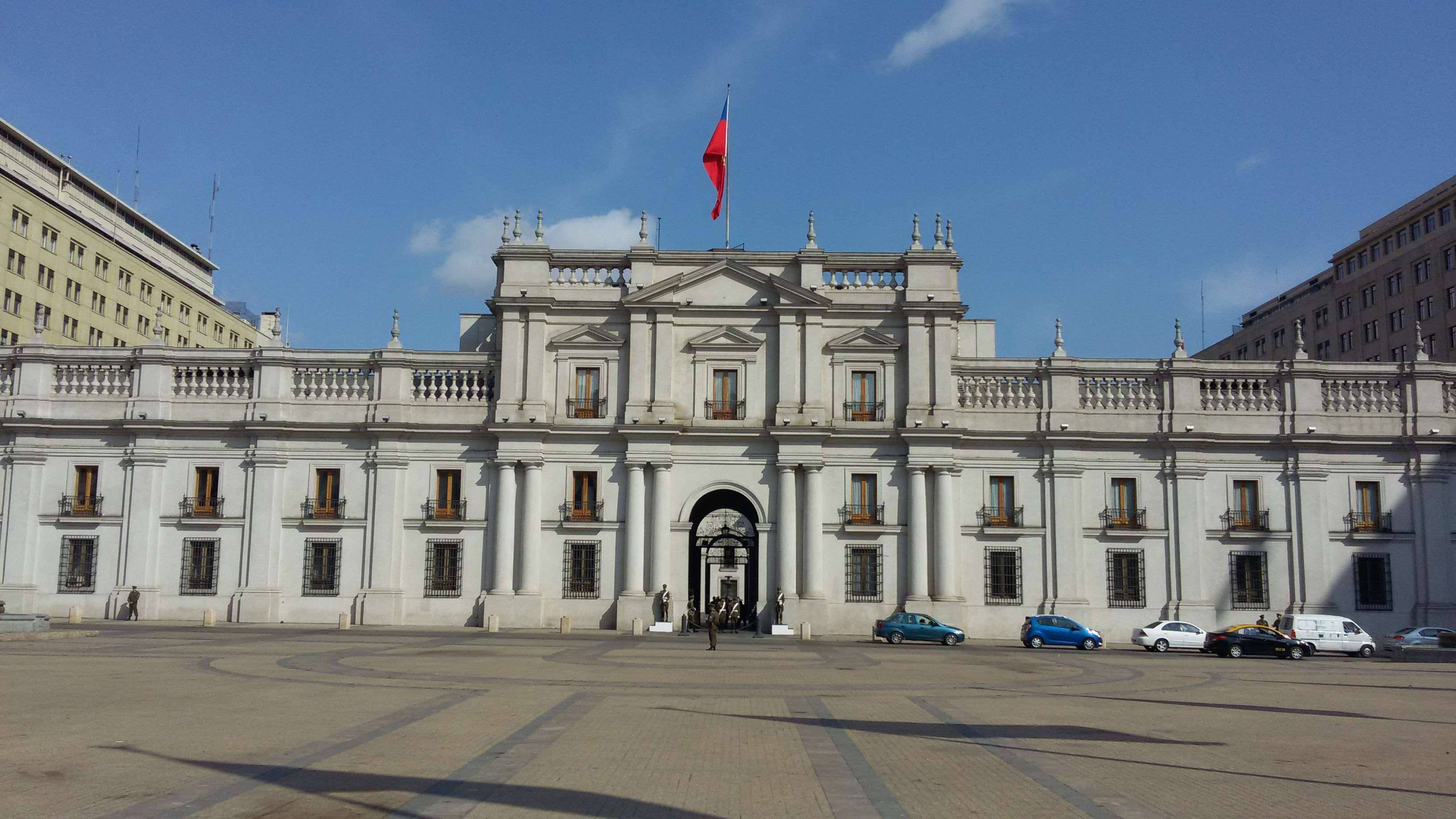 Chile 桌布 壁紙 — 一座宏偉的新古典主義白色政府大樓，頂部飄揚著智利國旗，背景是清澈的藍天，前方停有汽車。
