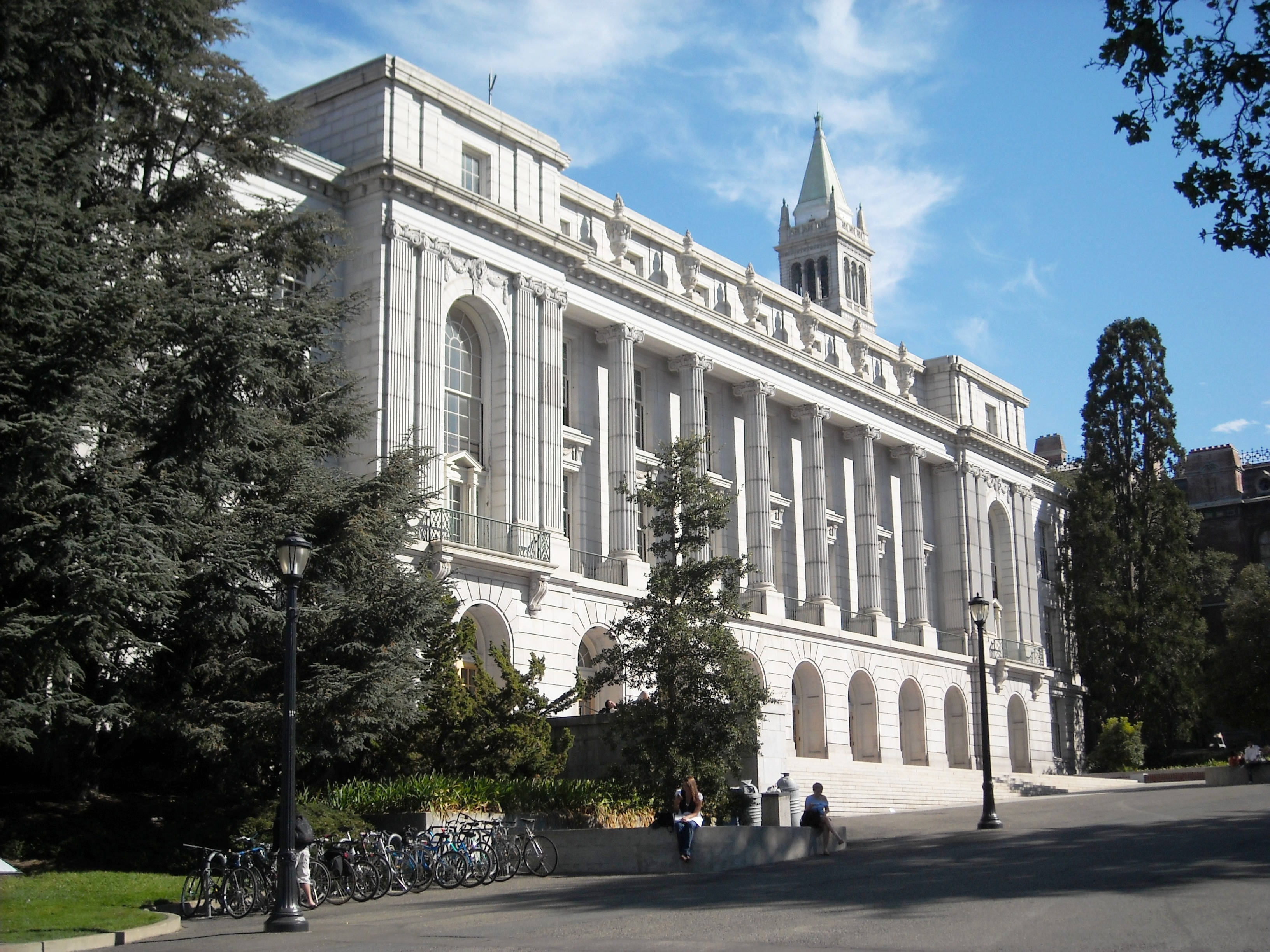 加州 桌布 壁紙 — 一座宏偉的新古典主義白色石造大學建築，擁有柱子和尖塔，在晴朗的藍天下，被高大的常綠樹木和學生環繞。
