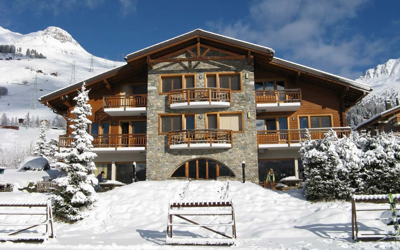 Ski Resort 桌布 壁紙 — 一座宏偉的木製阿爾卑斯山小屋，帶有石頭裝飾和積雪覆蓋的陽台，在明亮的藍天下，以白雪覆蓋的山脈為背景，傲然矗立。