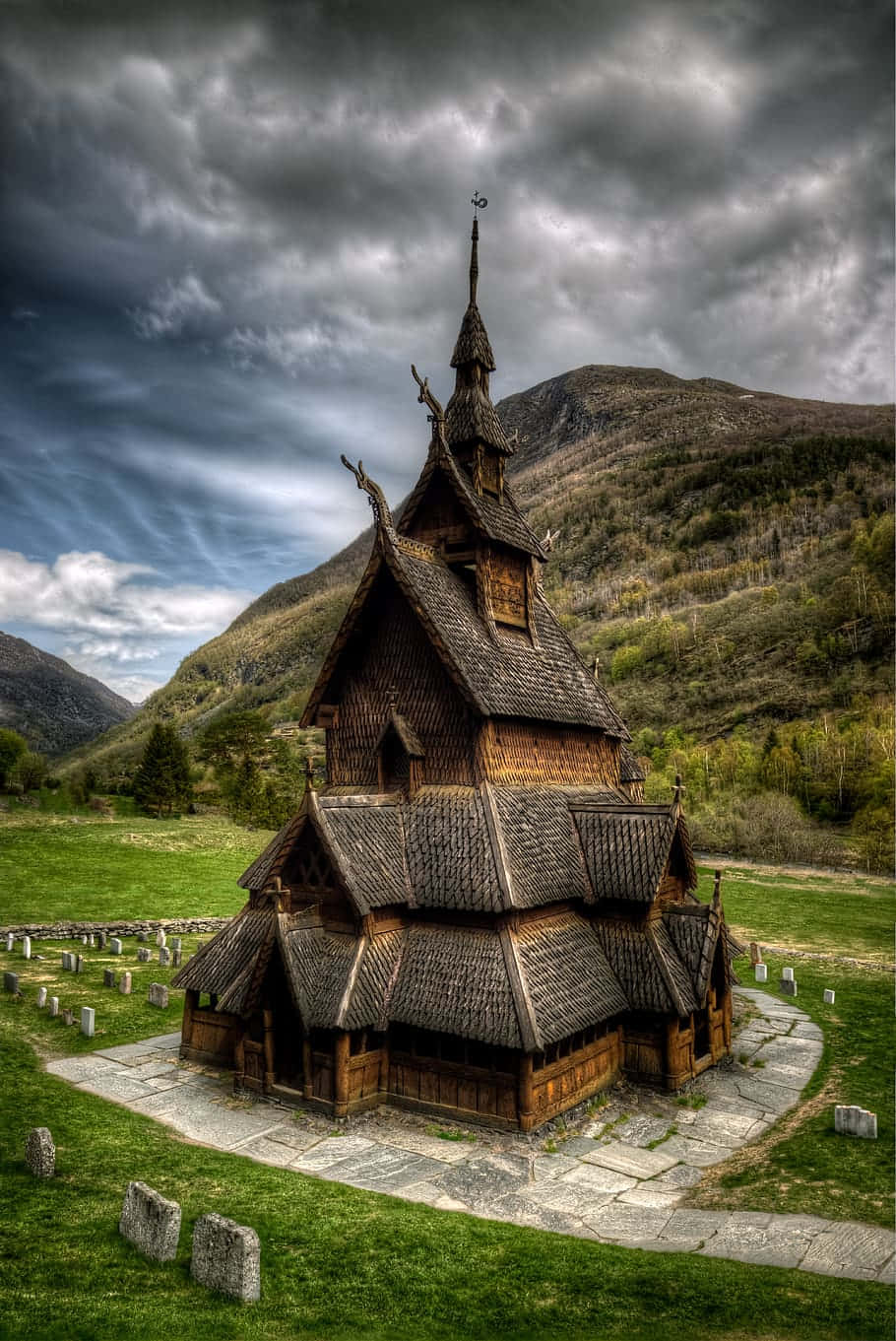 Norway 桌布 壁紙 — 一座宏偉的木造木板教堂，雕刻精細，莊嚴地矗立在長滿青草的山谷中，背景是戲劇性的暴風雨天空和山脈。
