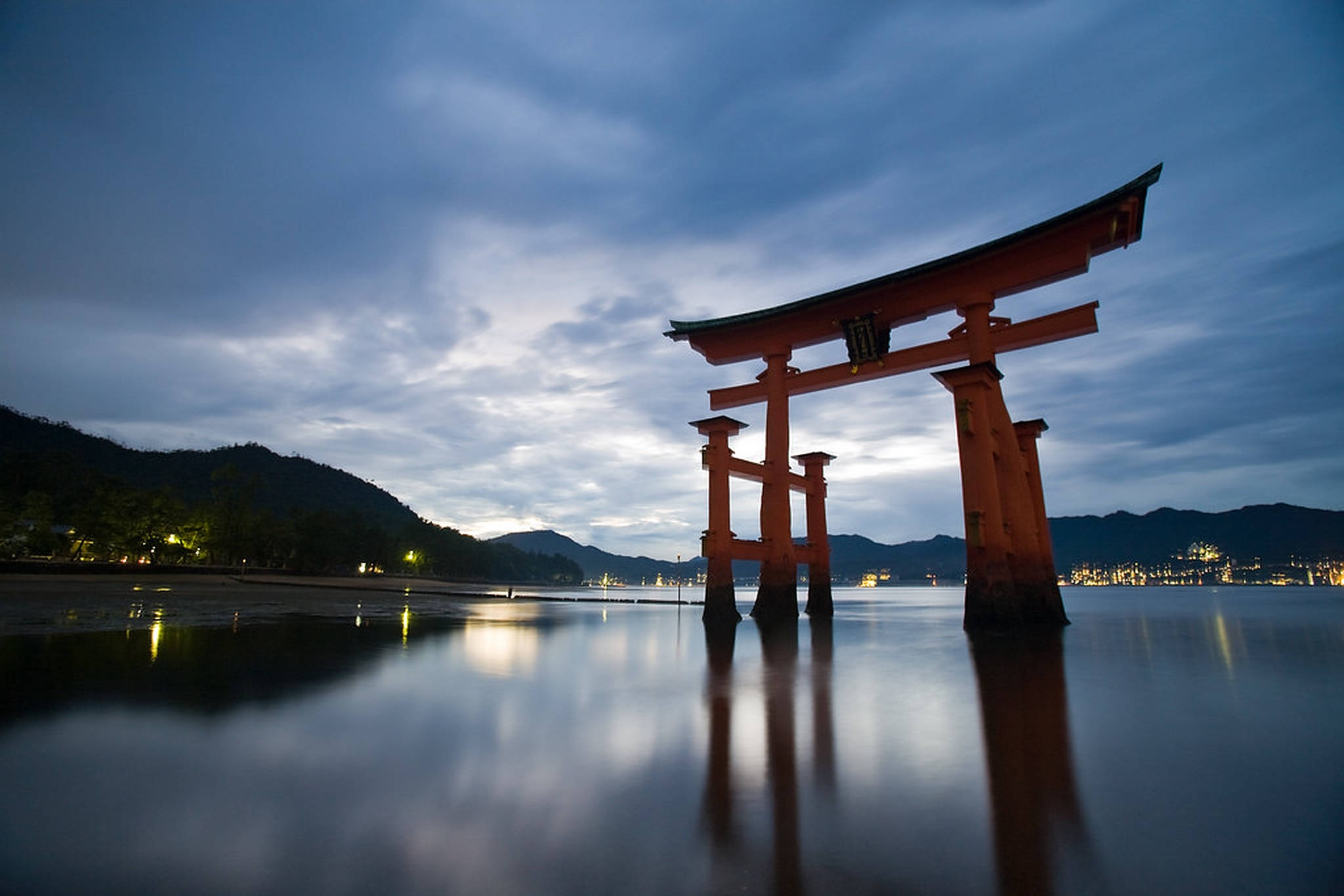 Torii 桌布 壁紙 — 一座宏偉的朱紅色鳥居矗立於黃昏時分的平靜水面上，遠山與城市燈光在陰沉天空下倒映其中。