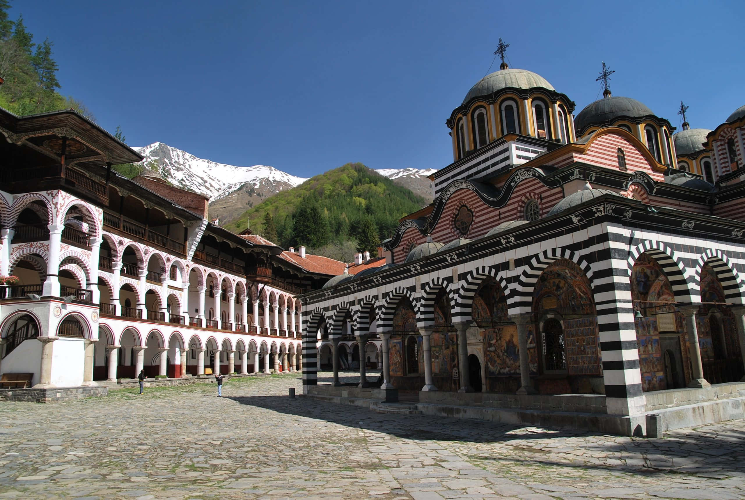Bulgaria 桌布 壁紙 — 一座宏偉的東正教修道院，擁有條紋拱門和圓頂，矗立在陽光明媚的庭院中，白雪覆蓋的山脈在晴朗的藍天下聳立於後。