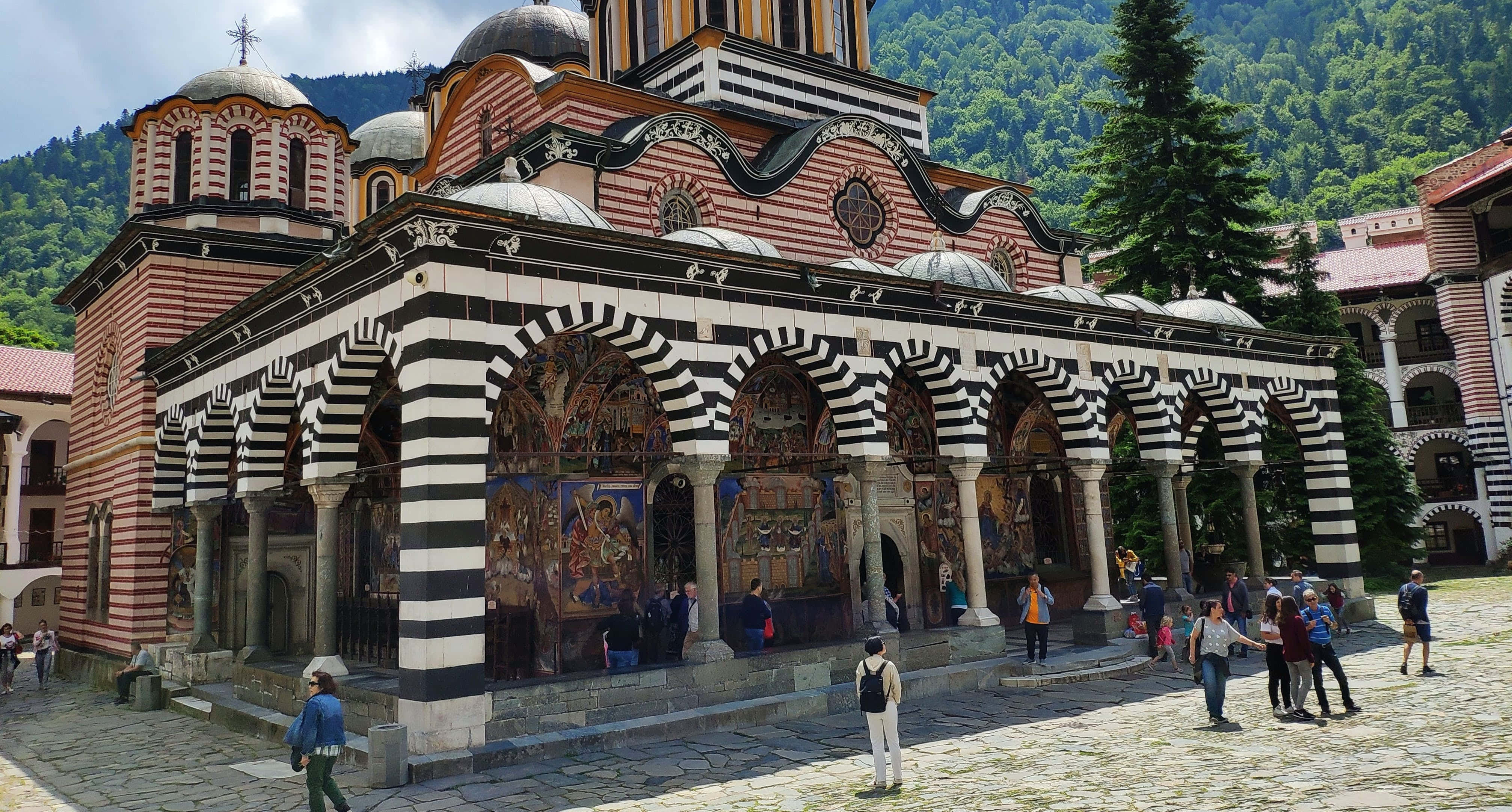 建築 桌布 壁紙 — 一座宏偉的條紋修道院，擁有拱形柱廊和圓頂，矗立在晴朗天空下鬱鬱蔥蔥的綠色山脈之中。