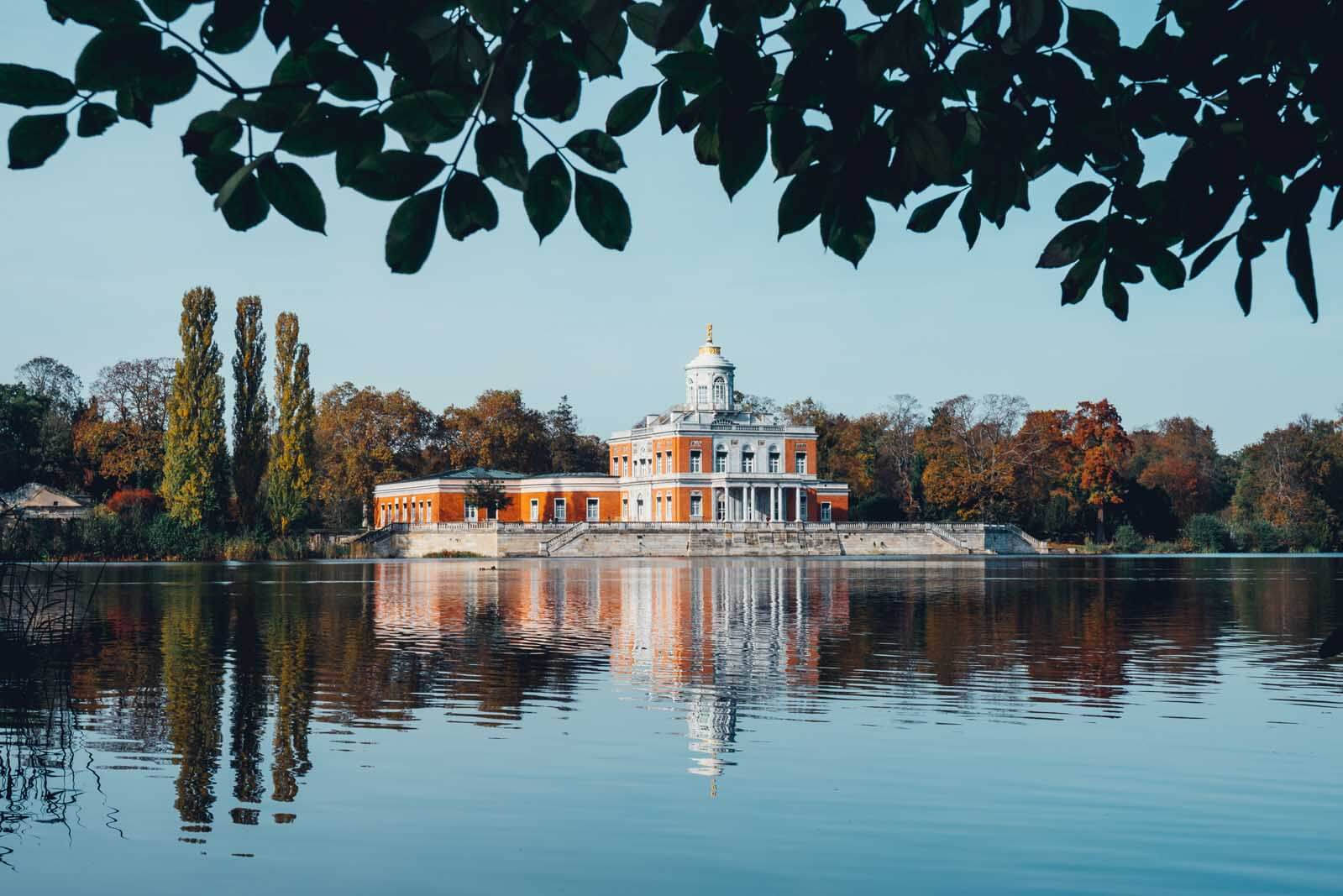 Potsdam 桌布 壁紙 — 一座宏偉的橙白色宮殿寧靜地倒映在平靜的湖面上，上方被深色的秋葉框住。