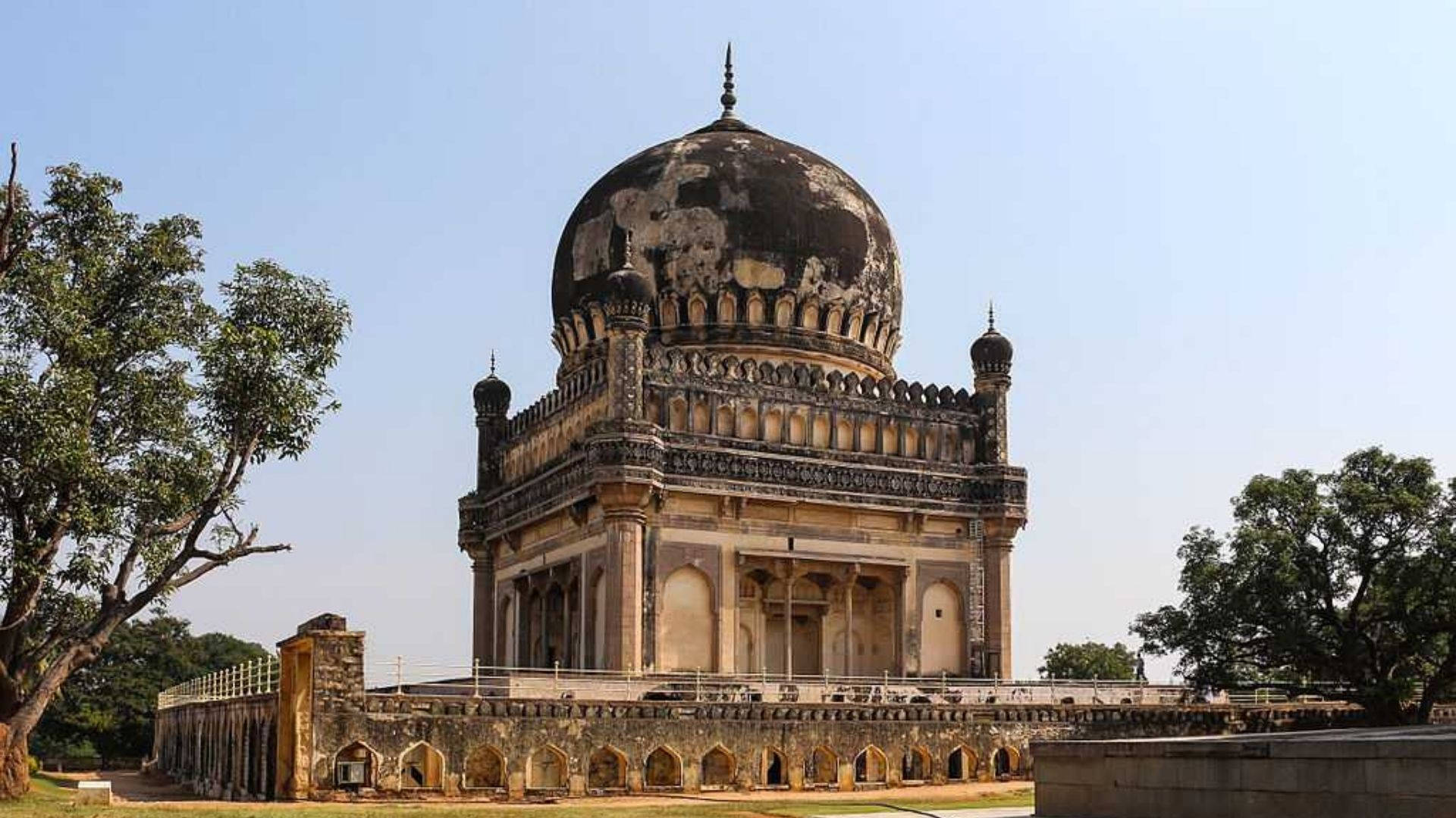 Hyderabad 桌布 壁紙 — 一座宏偉的歷史圓頂陵墓，帶有風化的石頭和精緻的雕刻，矗立在晴朗的藍天下，兩旁是茂密的綠樹。