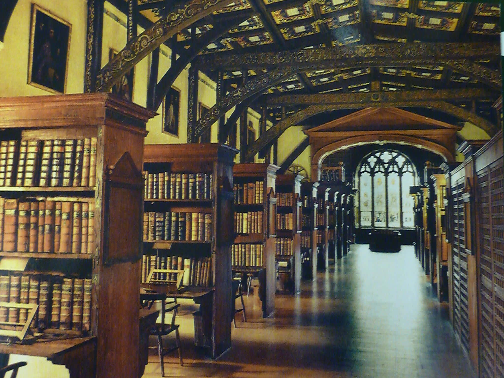 The Hogwarts Library 桌布 壁紙 — 一座宏偉的歷史圖書館，擁有高聳的木製書架、華麗的天花板橫樑，以及透過彩色玻璃窗過濾的溫暖金色光線。