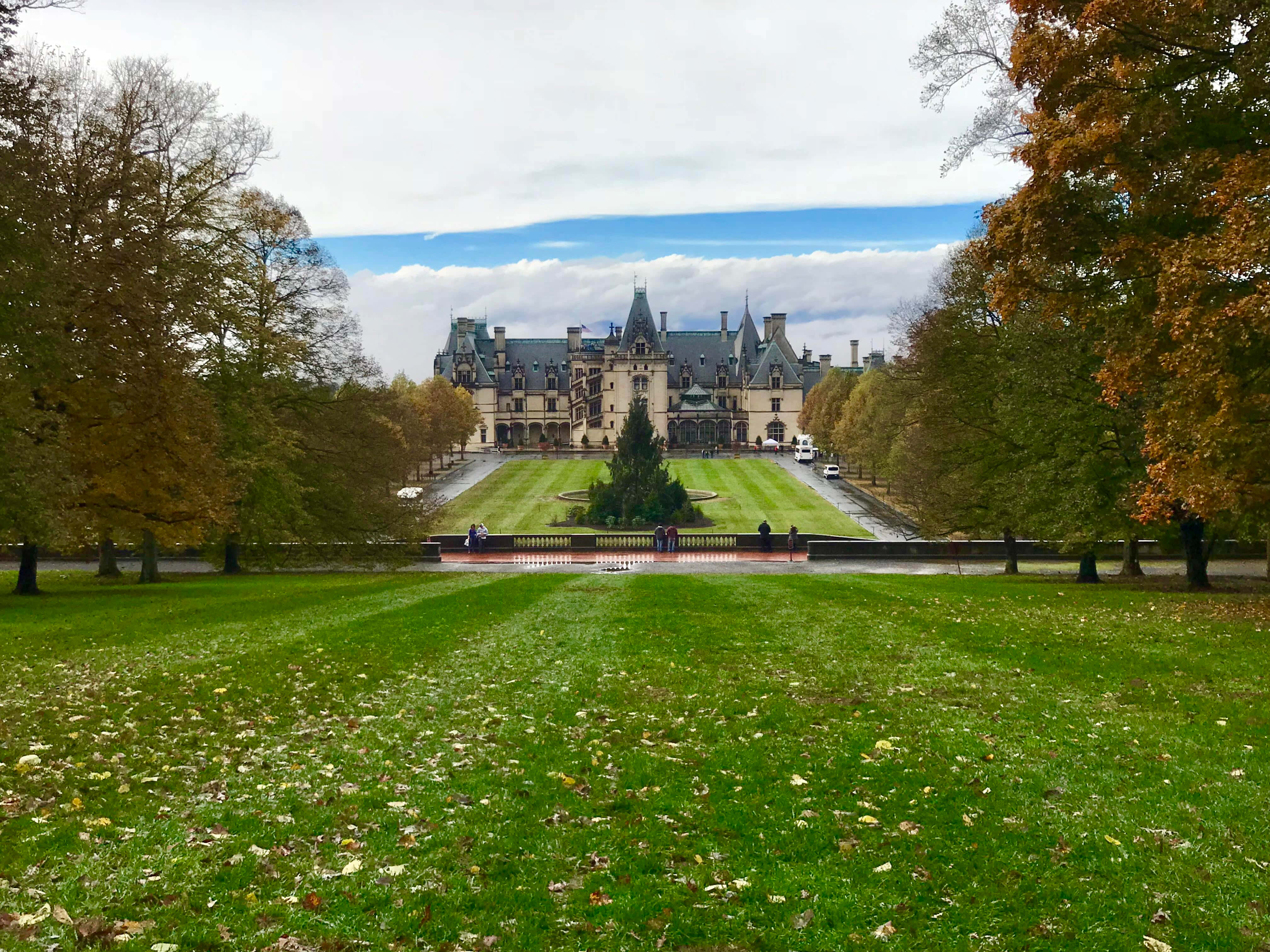 Biltmore Estate 桌布 壁紙 — 一座宏偉的歷史城堡，帶有塔樓和尖頂，矗立在長長修剪整齊的草坪盡頭，兩旁種滿了秋天的樹木，天空多雲。