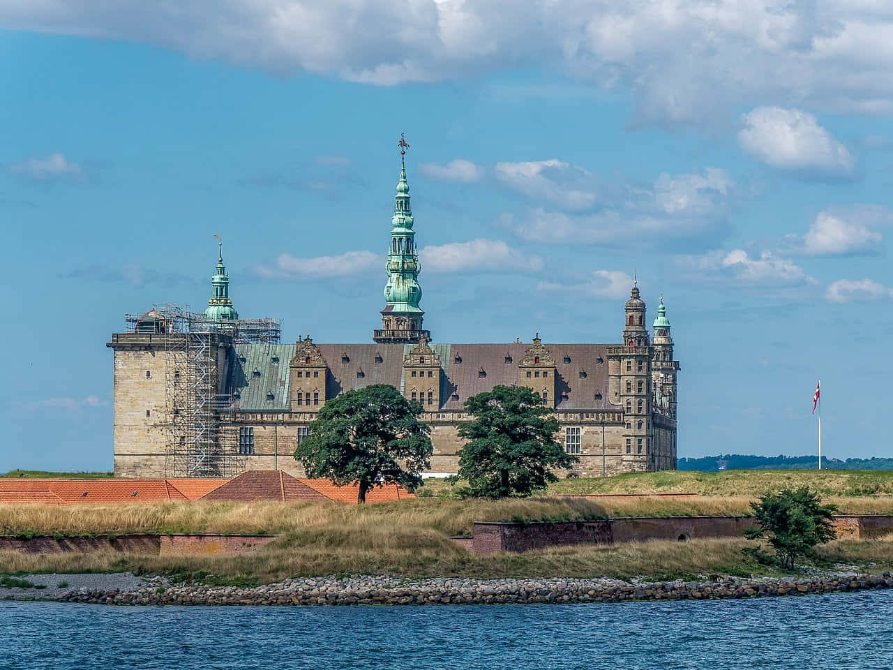 Denmark 桌布 壁紙 — 一座宏偉的歷史城堡，帶有綠色銅製尖頂，雄偉地矗立在一個長滿草的小島上，周圍是平靜的藍色海水，天空部分多雲。
