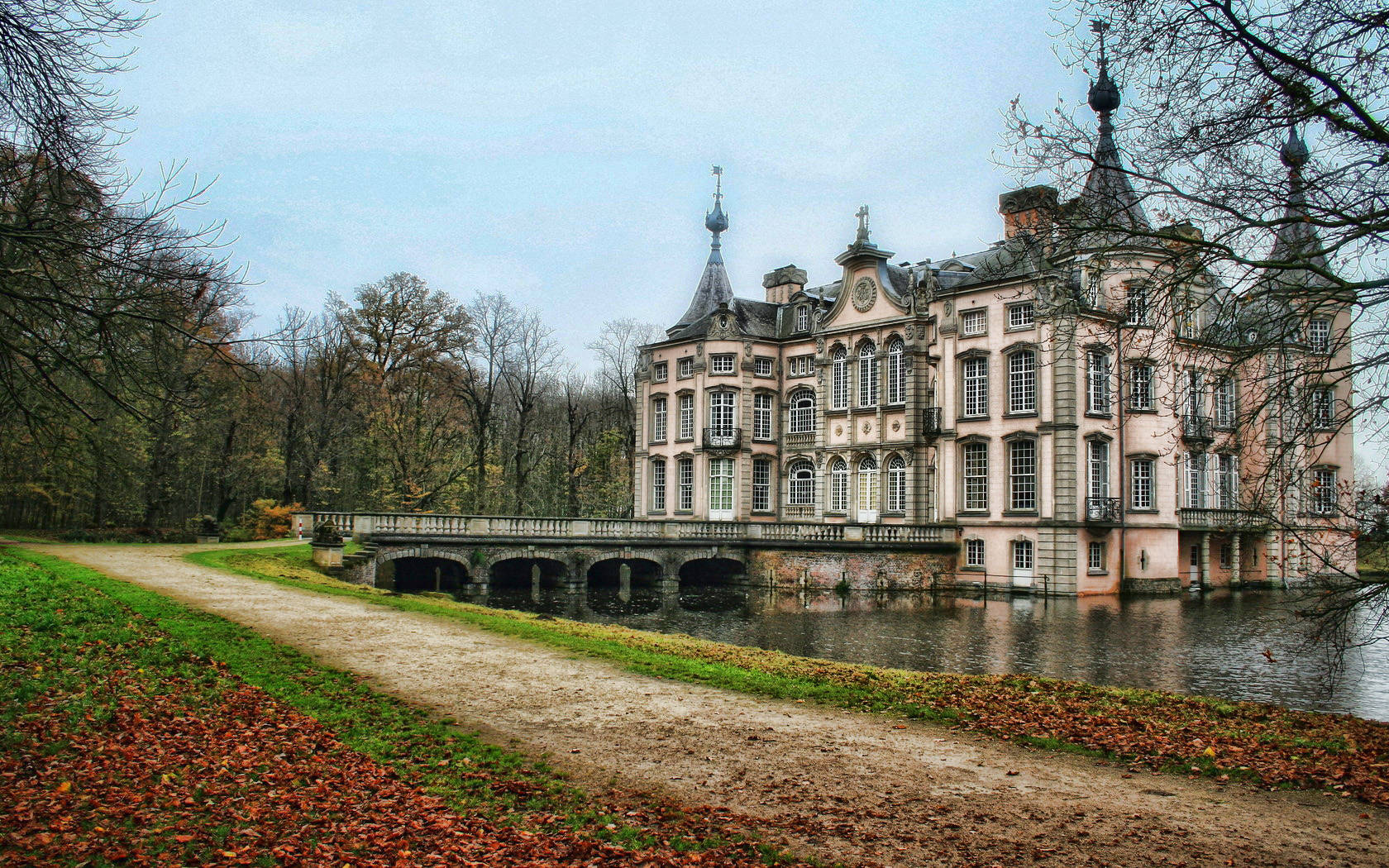 Belgium 桌布 壁紙 — 一座宏偉的歷史城堡，擁有華麗的塔樓，倒映在平靜的水面上，被秋天的樹木和一條落葉覆蓋的小徑環繞，天空多雲。