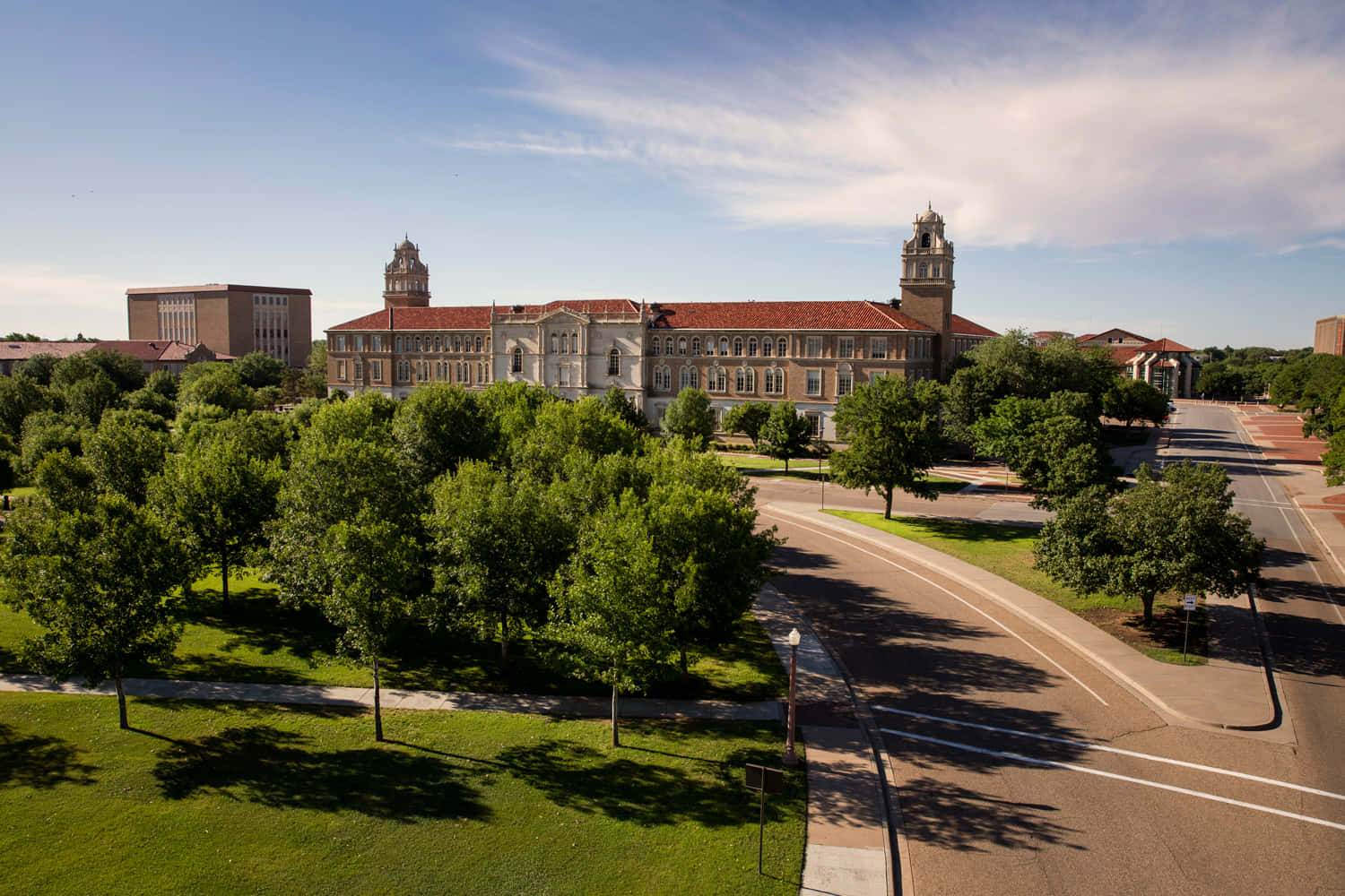 Texas Tech 桌布 壁紙 — 一座宏偉的歷史大學建築，擁有紅色瓦片屋頂和雙塔，矗立在茂密的綠樹之中，天空湛藍，雲朵輕盈。