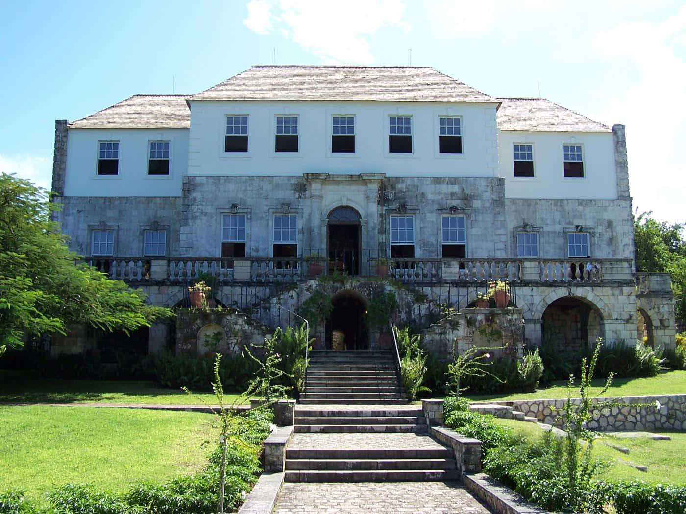 Jamaica 桌布 壁紙 — 一座宏偉的歷史大宅，白色上層牆壁和石質下層，在明亮的藍天下，由寬闊的石階進入，兩旁是茂密的綠色草坪和樹木。