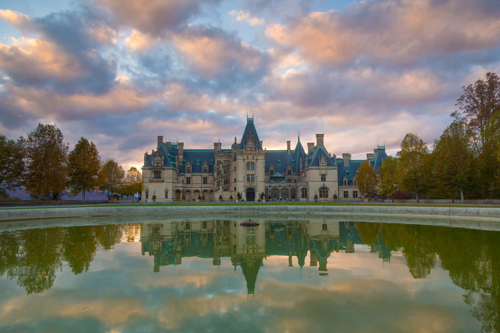 Biltmore Estate 桌布 壁紙 — 一座宏偉的歷史大宅，藍色屋頂在戲劇性的日落天空下完美倒映在平靜的池塘中。