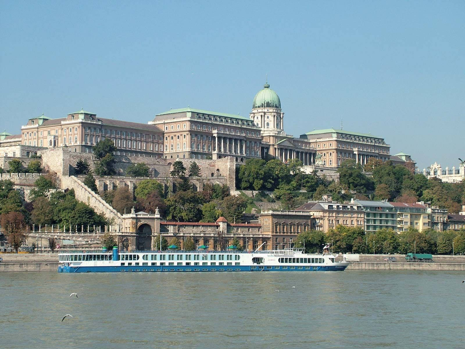 Hungary 桌布 壁紙 — 一座宏偉的歷史宮殿坐落在山坡上，俯瞰著一條寬闊的河流，一艘藍白相間的遊輪在清澈的藍天下航行。
