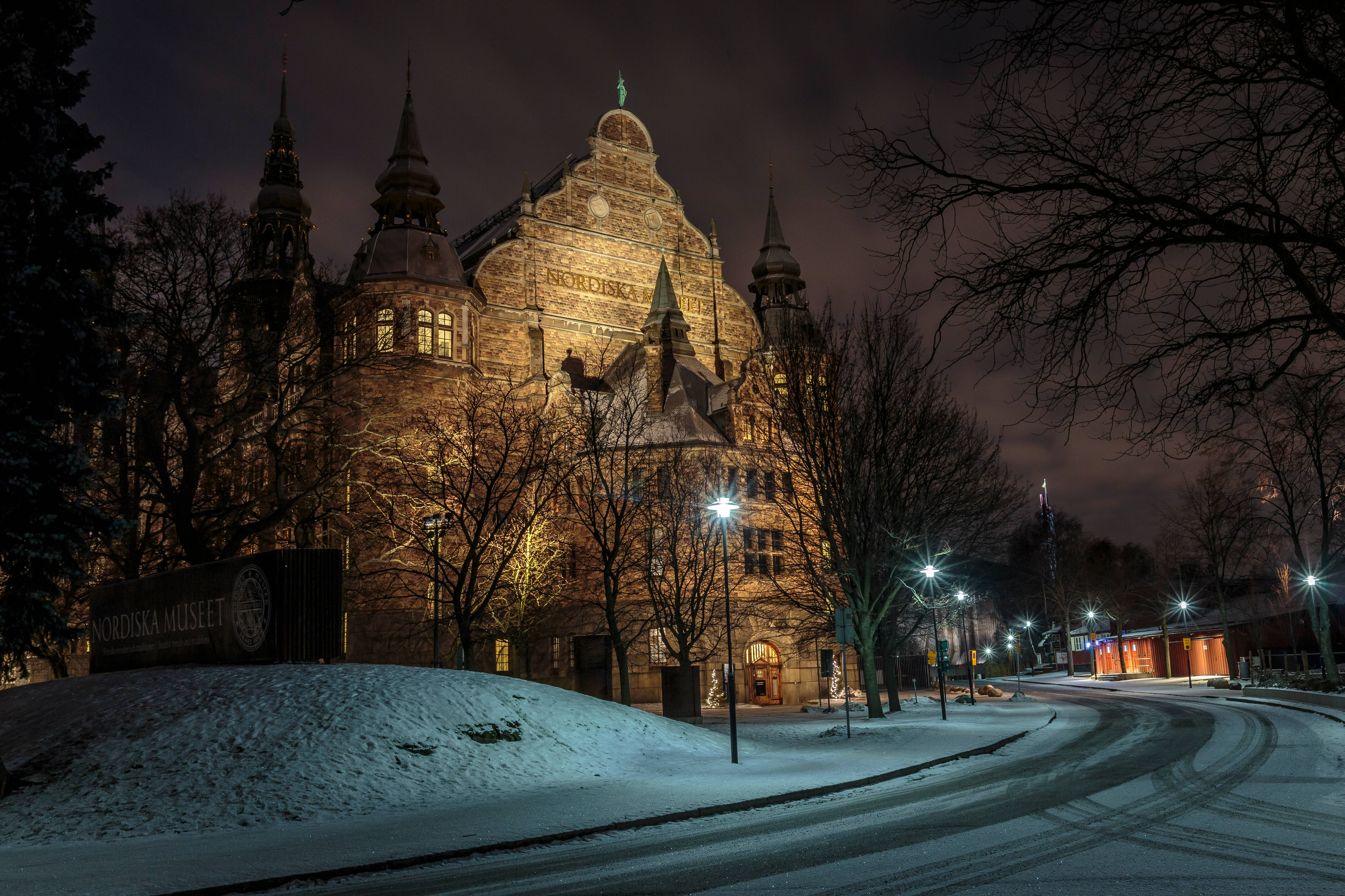 Sweden 桌布 壁紙 — 一座宏偉的歷史建築，帶有尖頂和溫暖的窗戶，在街燈下閃耀，背景是雪夜天空和光禿的樹木。