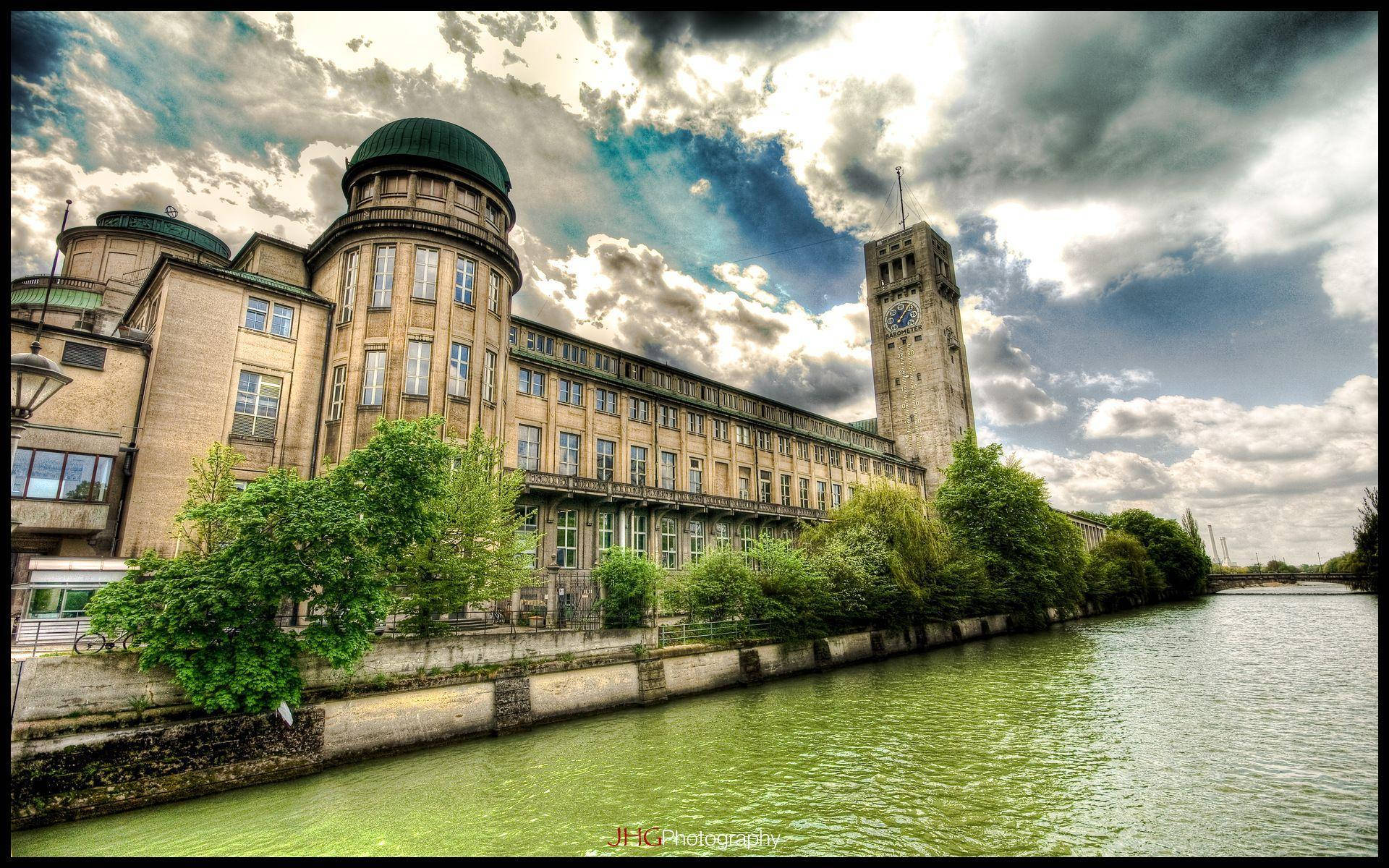 Munich 桌布 壁紙 — 一座宏偉的歷史建築，帶有鐘樓，矗立在平靜的綠色運河旁，天空戲劇性地多雲。
