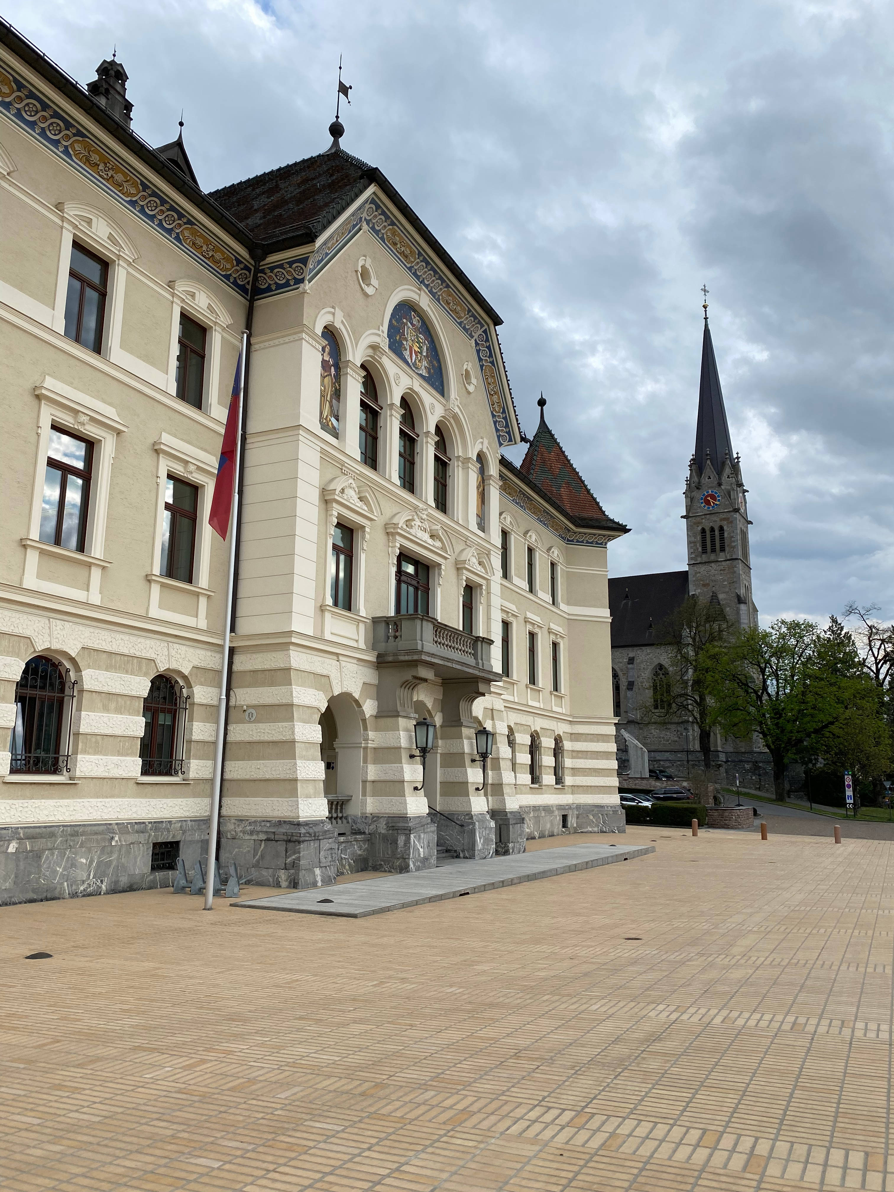 Liechtenstein 桌布 壁紙 — 一座宏偉的歷史建築，擁有華麗的外牆和旗杆，矗立在教堂尖頂旁，天空多雲。