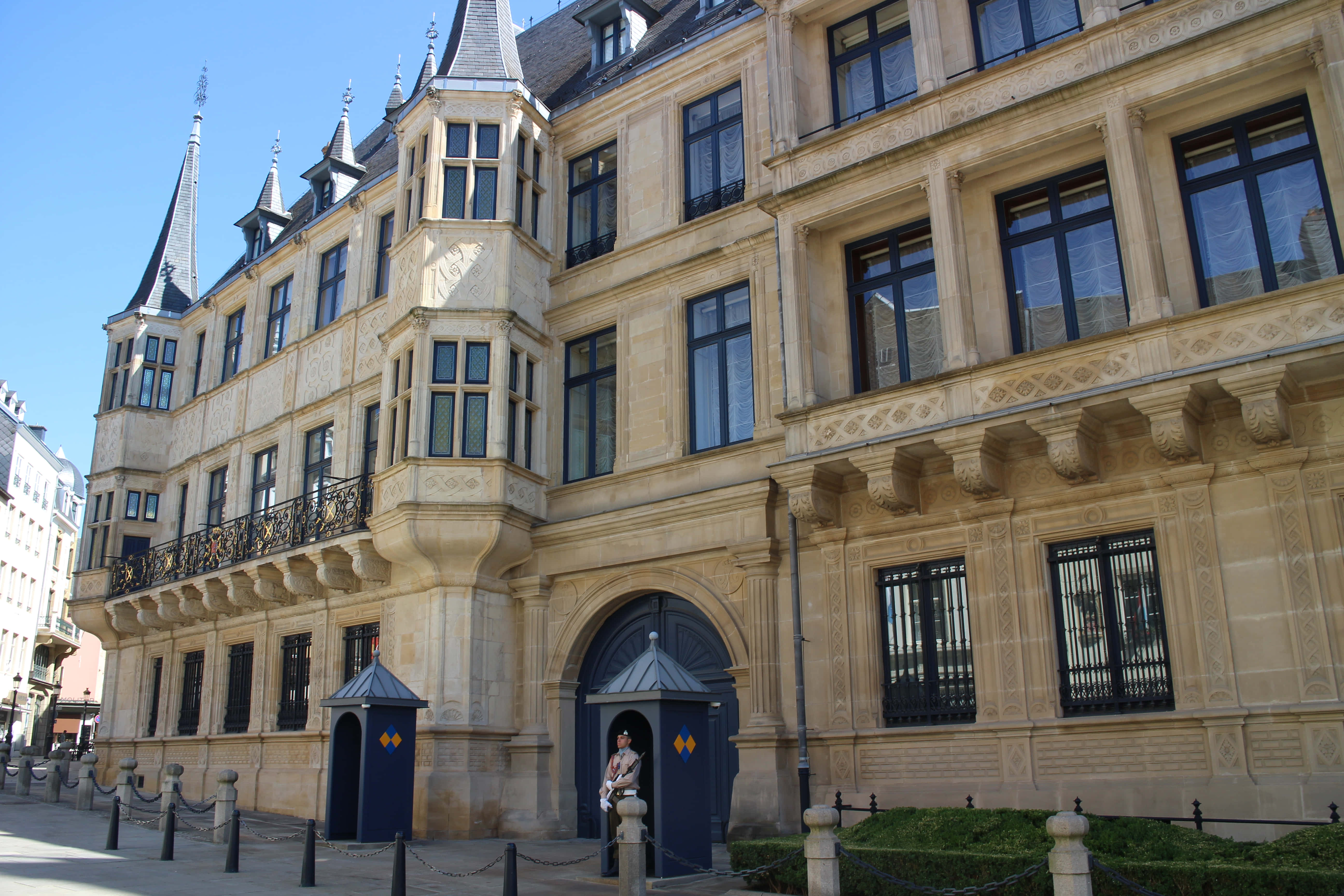 Palace 桌布 壁紙 — 一座宏偉的歷史建築，擁有華麗的石砌外牆、黑色窗戶和尖塔，在晴朗的藍天下矗立，入口處有警衛。