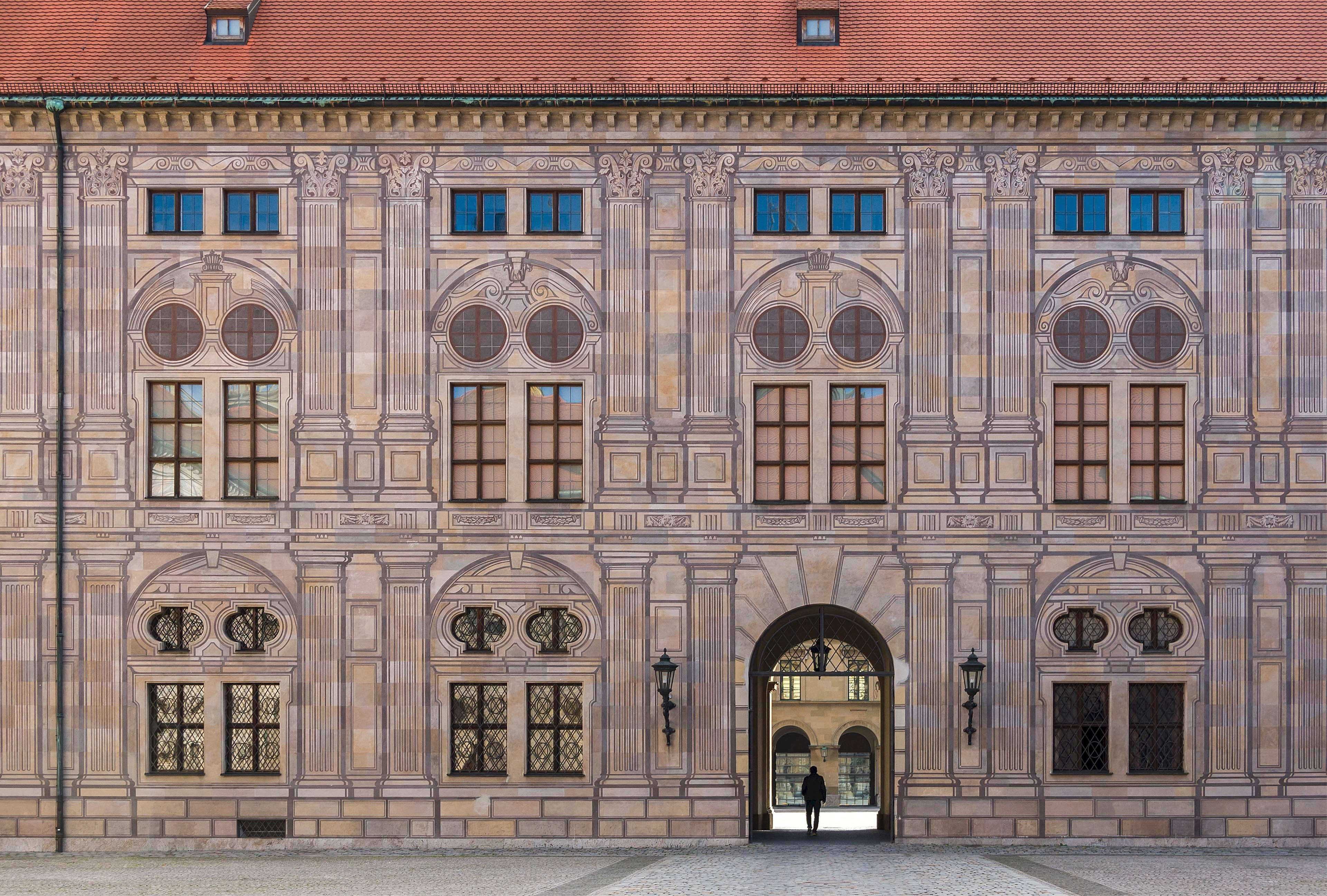 Munich 桌布 壁紙 — 一座宏偉的歷史建築，擁有華麗的石砌外牆和紅色屋頂，中央有一個拱形大門，一個孤獨的身影正穿過其中。