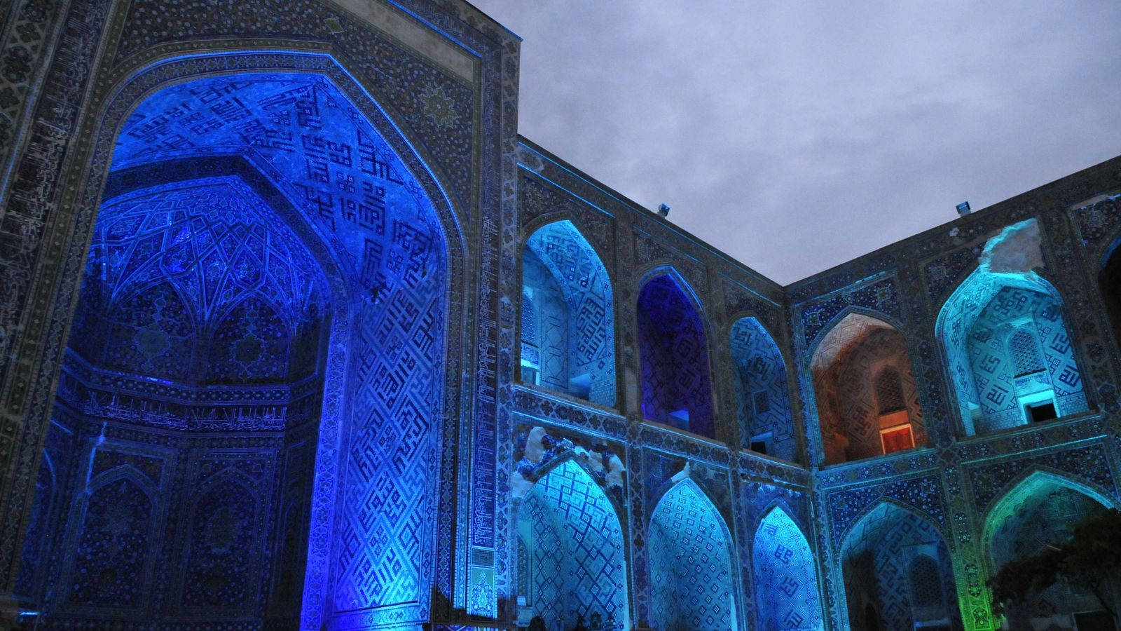 Uzbekistan 桌布 壁紙 — 一座宏偉的歷史建築，擁有複雜的藍色瓷磚拱門，在多雲的暮色天空下被照亮。