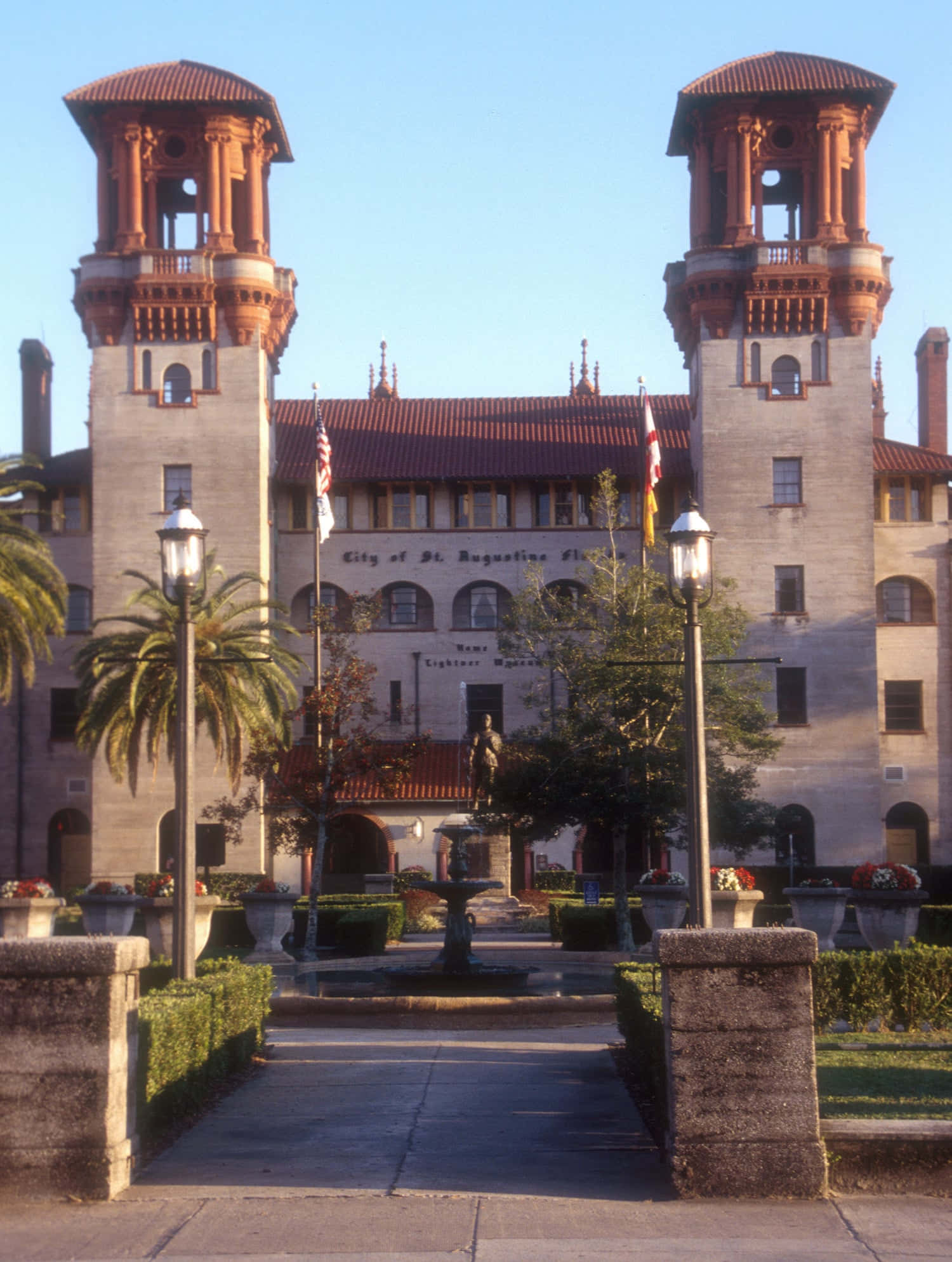 Saint Augustine Florida 桌布 壁紙 — 一座宏偉的歷史建築，擁有雙塔和紅瓦屋頂，矗立在清澈的藍天下，以棕櫚樹和中央噴泉為框景。