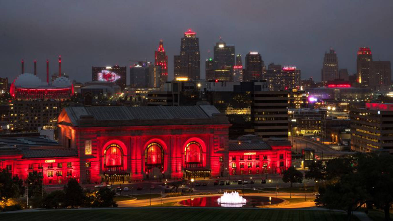 Kansas City 桌布 壁紙 — 一座宏偉的歷史建築沐浴在紅光中，矗立在黃昏的城市天際線前，下方有燈火通明的摩天大樓和發光的噴泉。