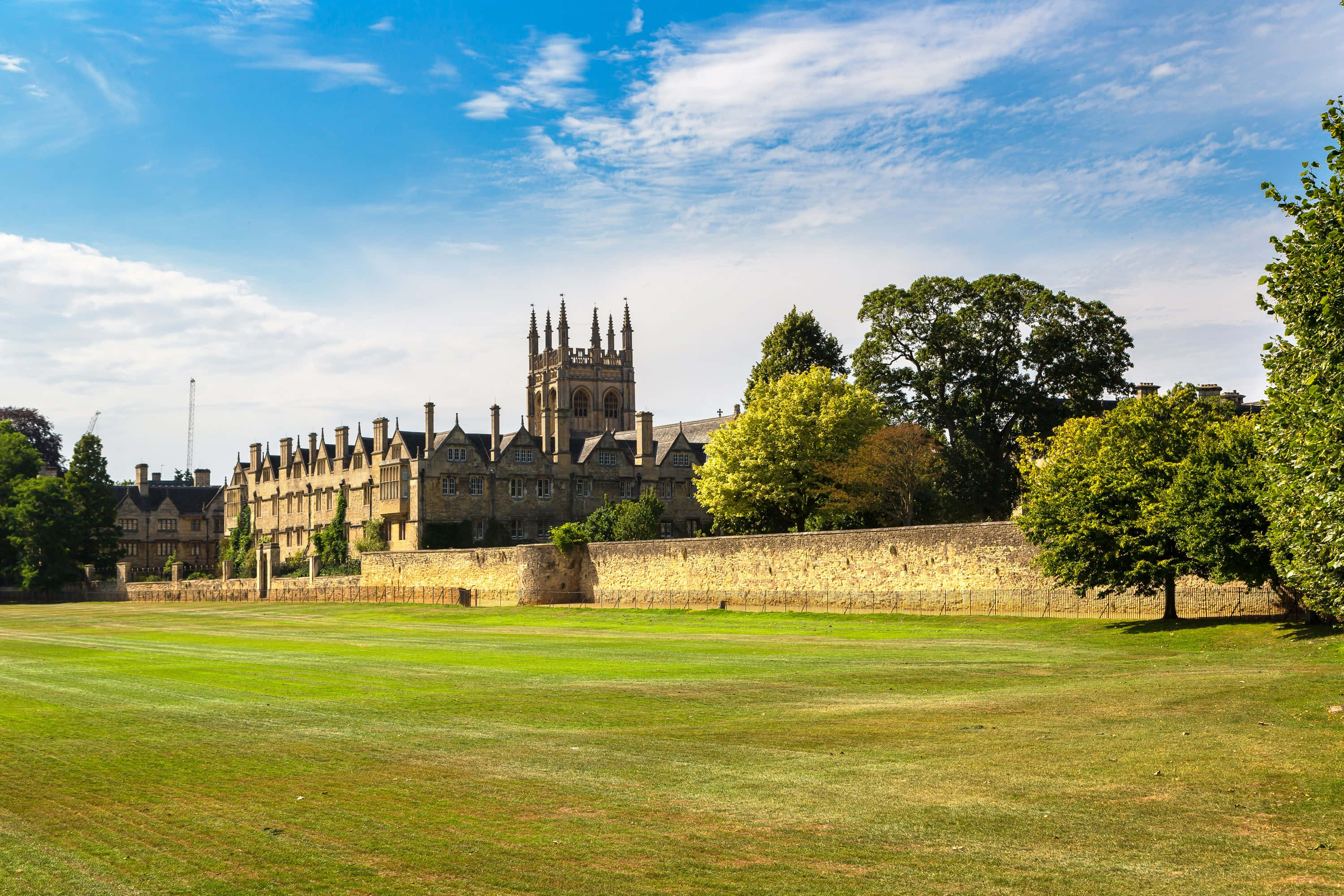 England 桌布 壁紙 — 一座宏偉的歷史悠久大學學院，帶有哥德式尖塔，矗立在石牆和茂密綠草之後，在明亮的藍天和零星雲朵下。