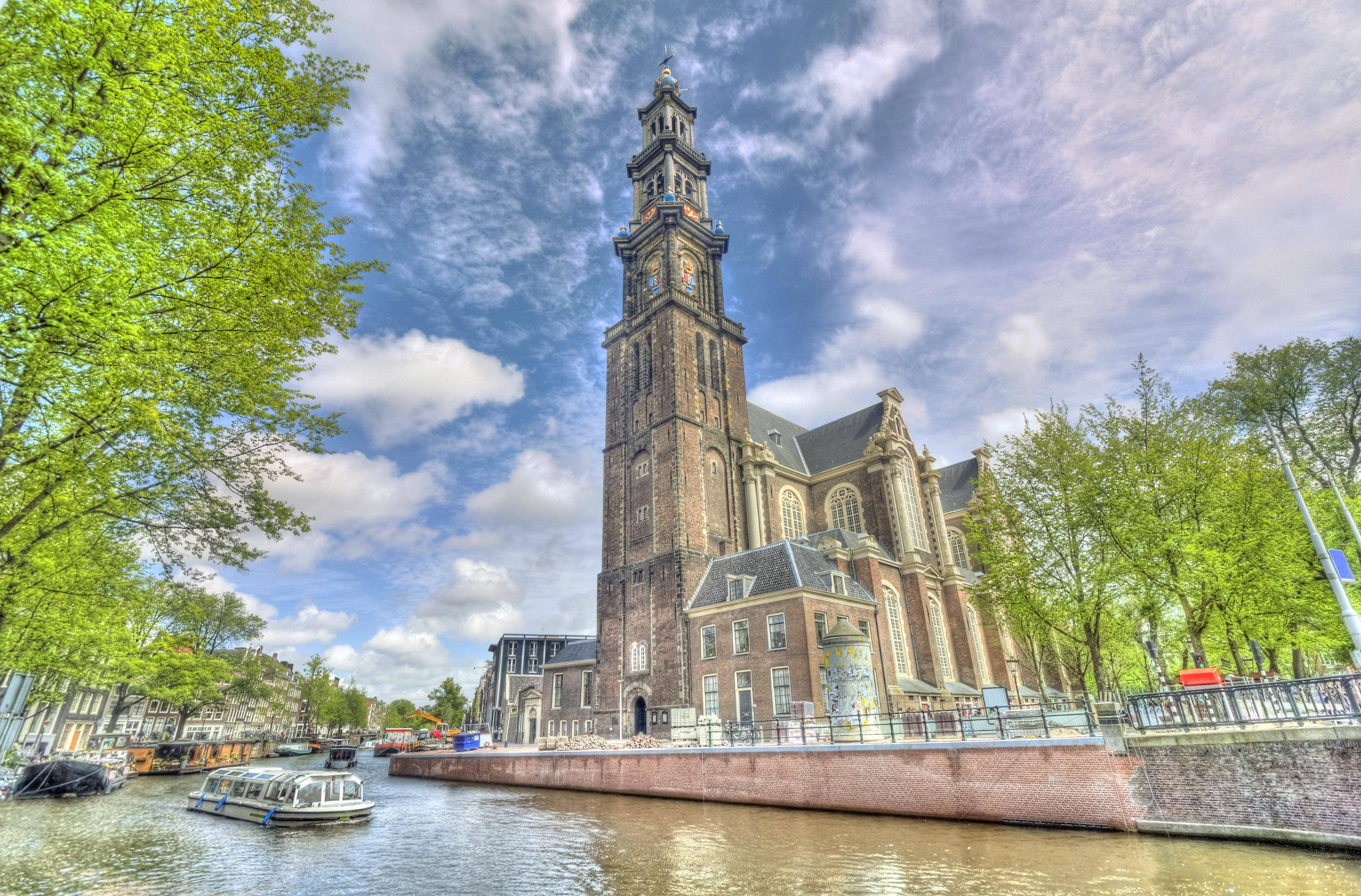 Amsterdam 桌布 壁紙 — 一座宏偉的歷史教堂，擁有高塔，矗立在運河旁，運河上有船隻，天空湛藍，點綴著蓬鬆的白雲。