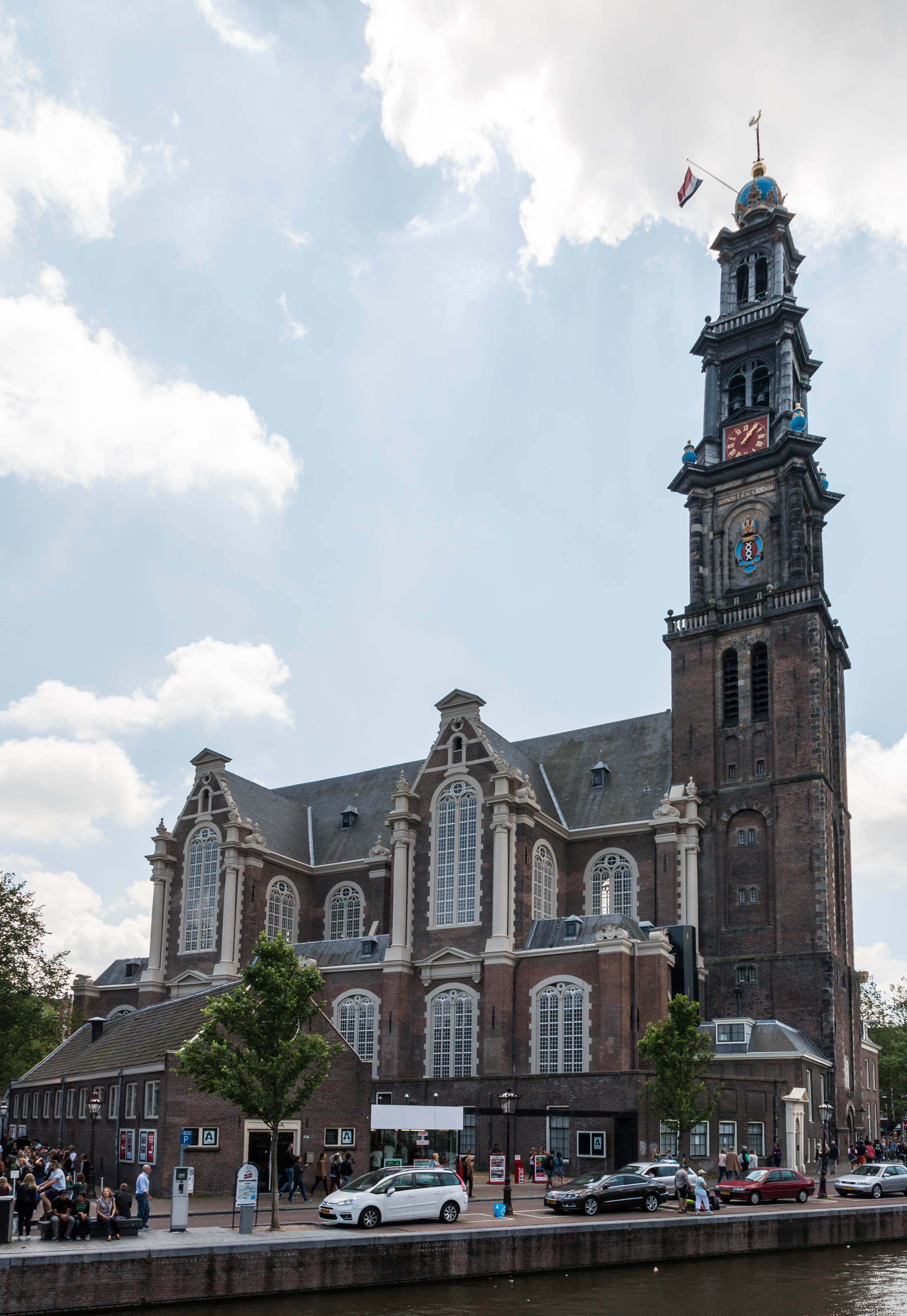 Amsterdam 桌布 壁紙 — 一座宏偉的歷史教堂，擁有高高的鐘樓，矗立在運河旁，天空多雲，附近有汽車和行人。