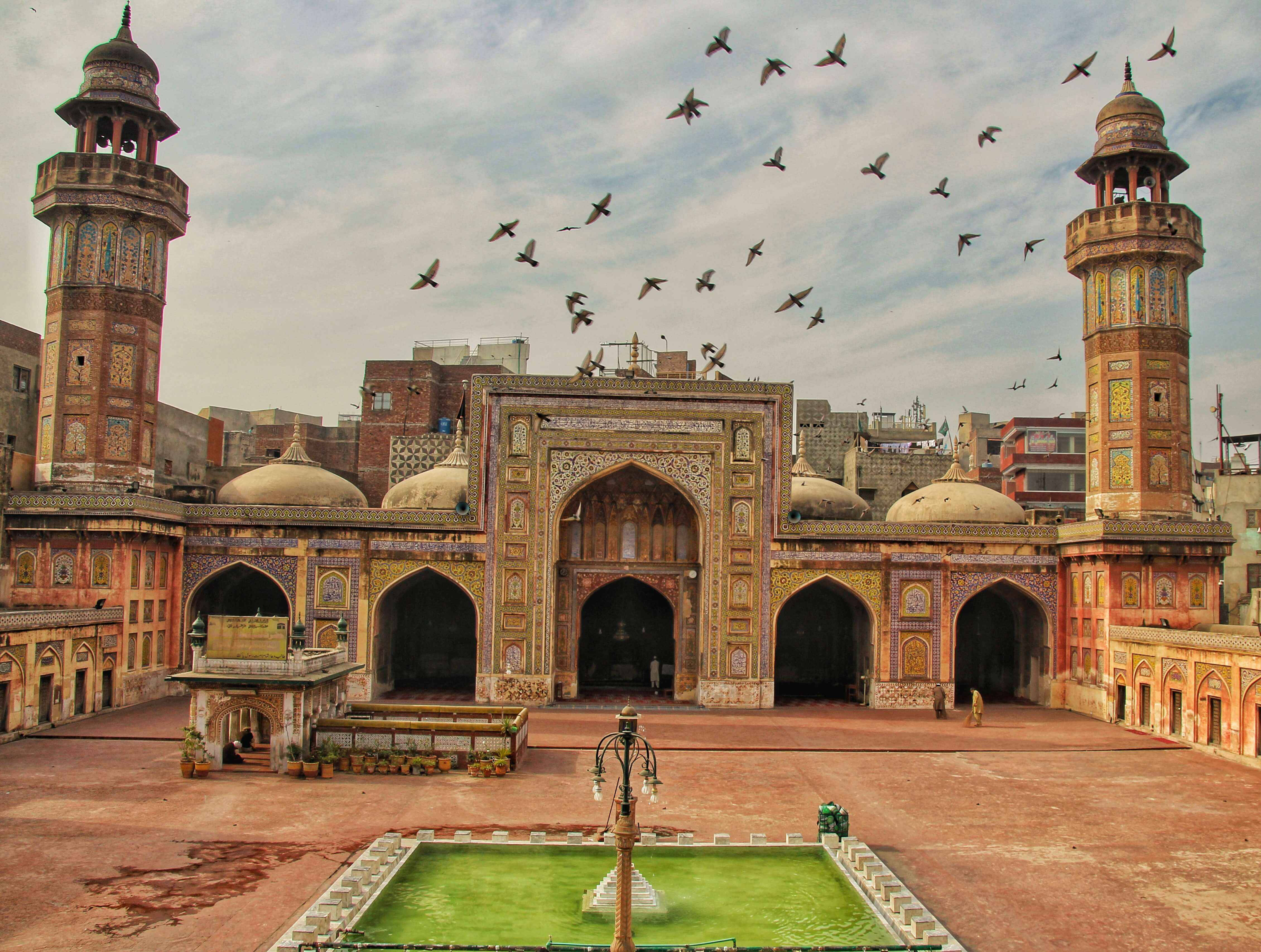Lahore 桌布 壁紙 — 一座宏偉的歷史清真寺，擁有華麗的圓頂和宣禮塔，矗立在多雲的天空下，鳥兒在頭頂翱翔，綠色噴泉在庭院中閃閃發光。