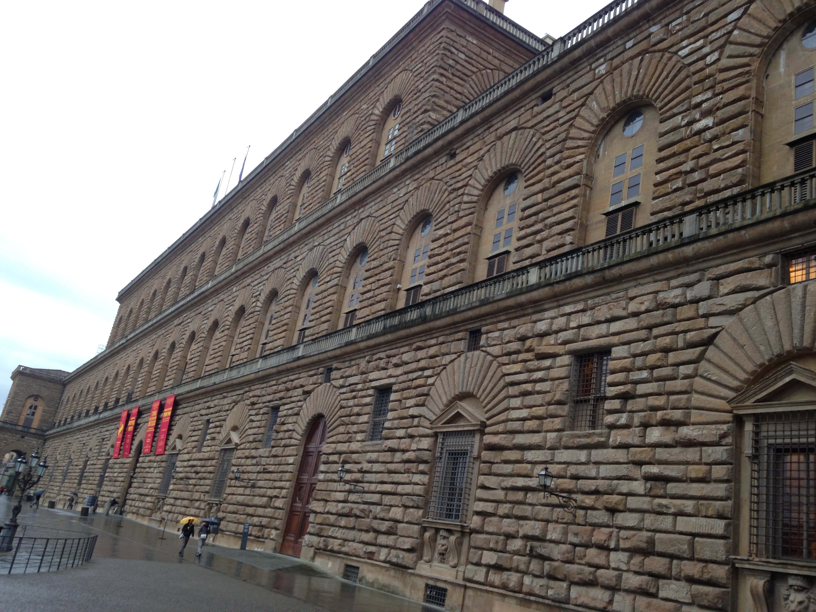 Building 桌布 壁紙 — 一座宏偉的歷史石建築，擁有拱形窗戶和陽台，天空陰沉，行人走在潮濕的路面上。