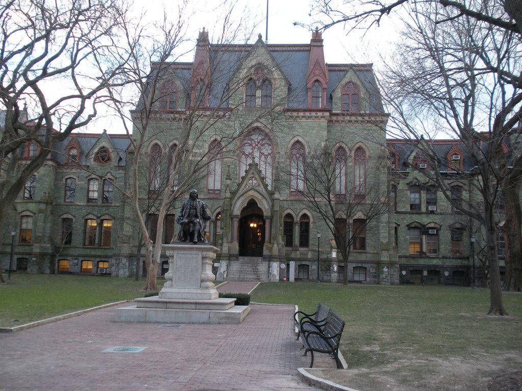 University Of Pennsylvania 桌布 壁紙 — 一座宏偉的歷史石砌哥德式建築，矗立在光禿的樹木和中央雕像後方，位於寧靜的校園庭院中。