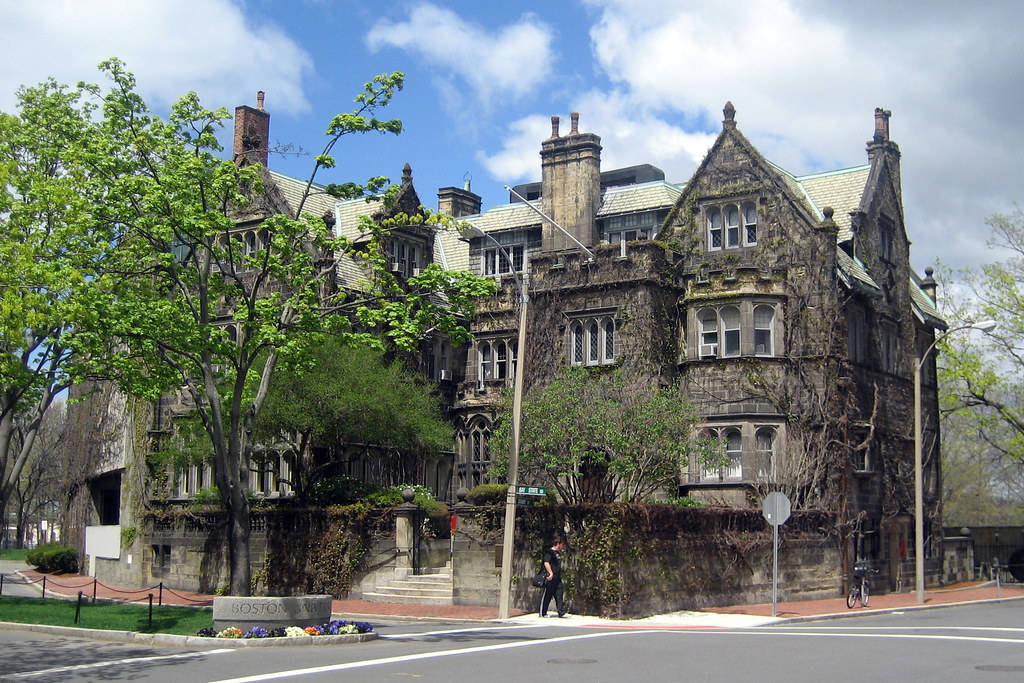 Building 桌布 壁紙 — 一座宏偉的歷史石造建築，牆壁爬滿常春藤，有多個煙囪，矗立在部分多雲的天空下，附近有茂密的綠樹。