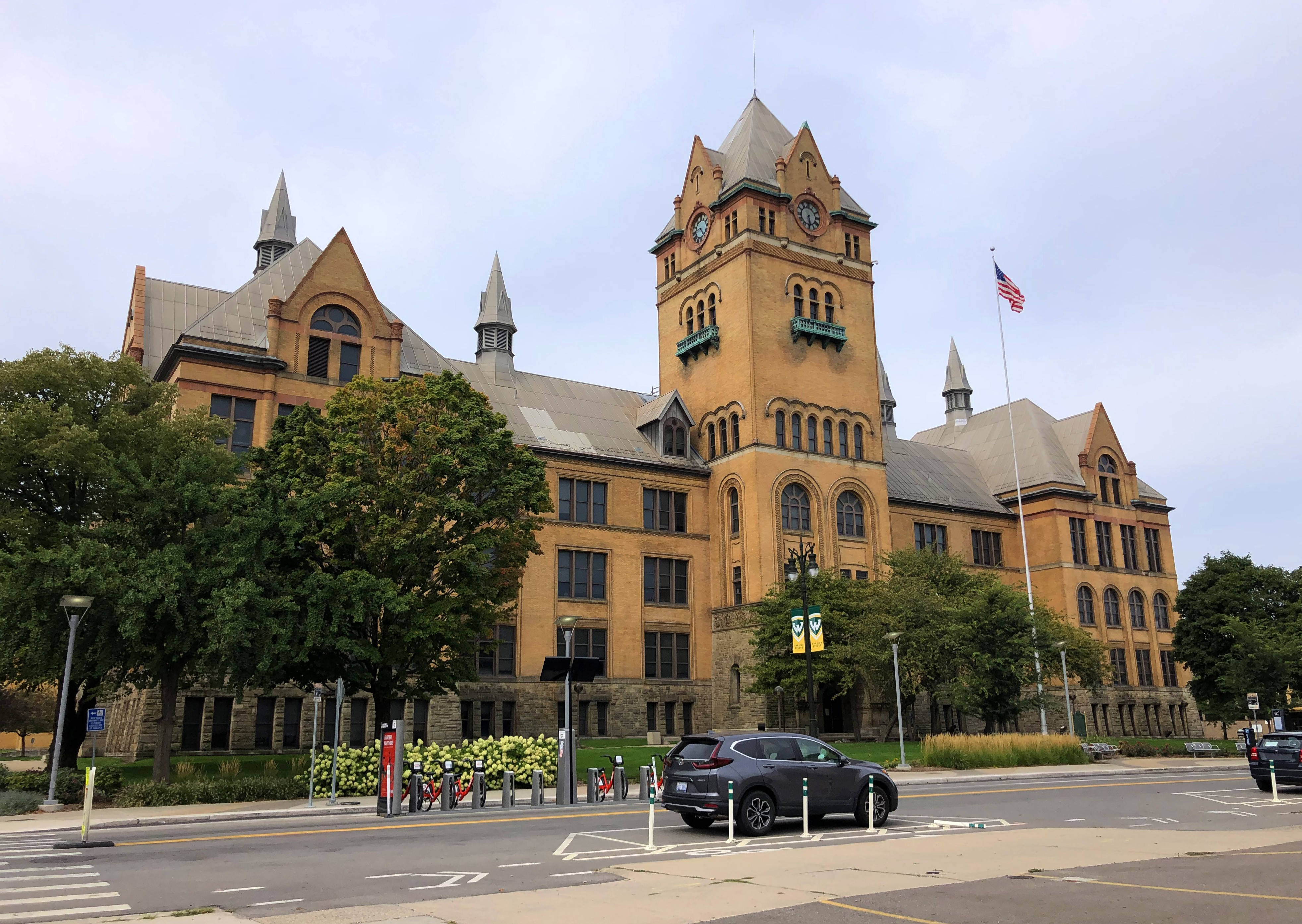 Building 桌布 壁紙 — 一座宏偉的歷史砂岩建築，帶有鐘樓和美國國旗，矗立在陰天之下，前面有樹木和一輛停著的汽車。