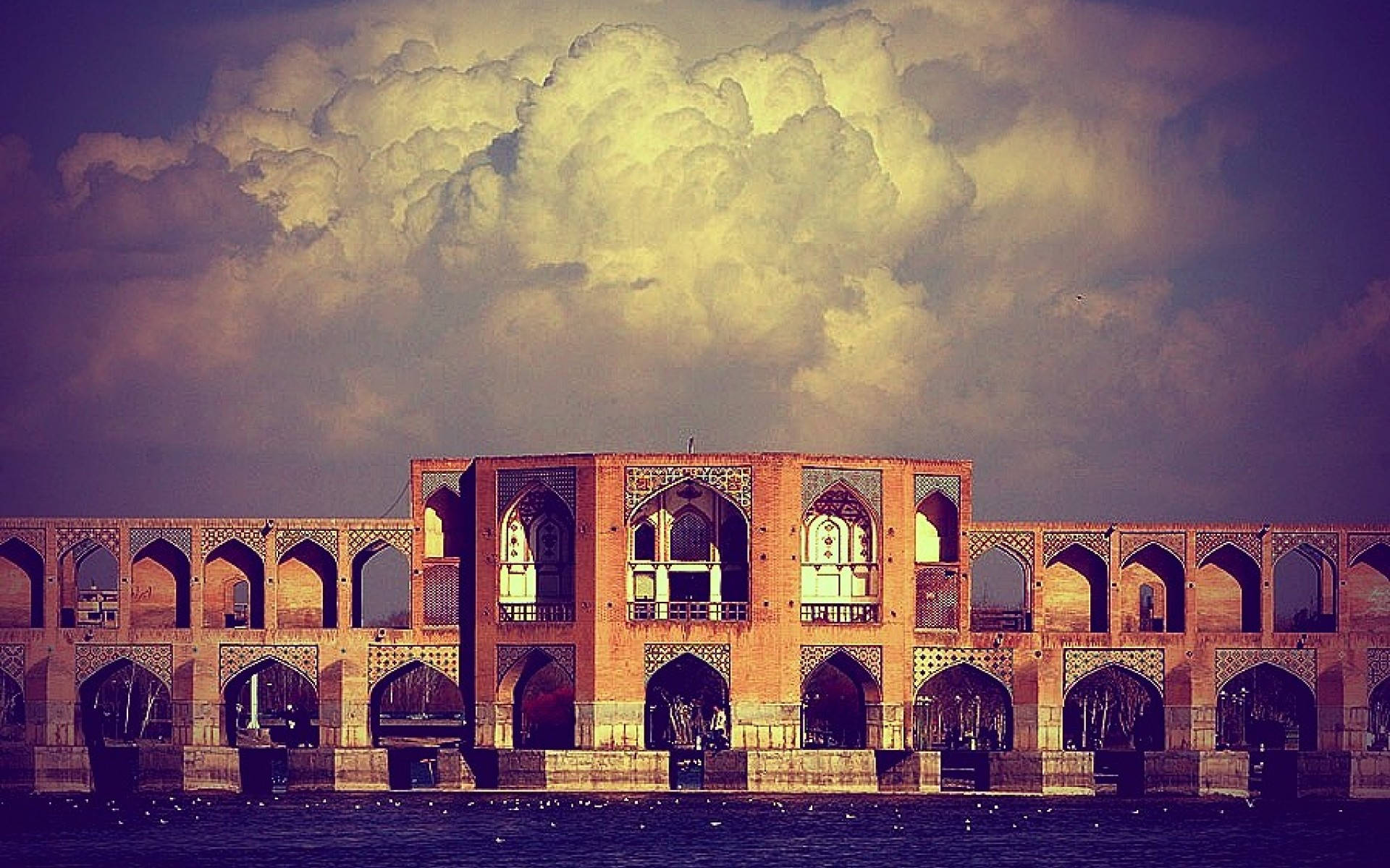 Tehran 桌布 壁紙 — 一座宏偉的歷史磚橋，帶有拱形開口，在戲劇性的多雲天空下橫跨平靜的河流。